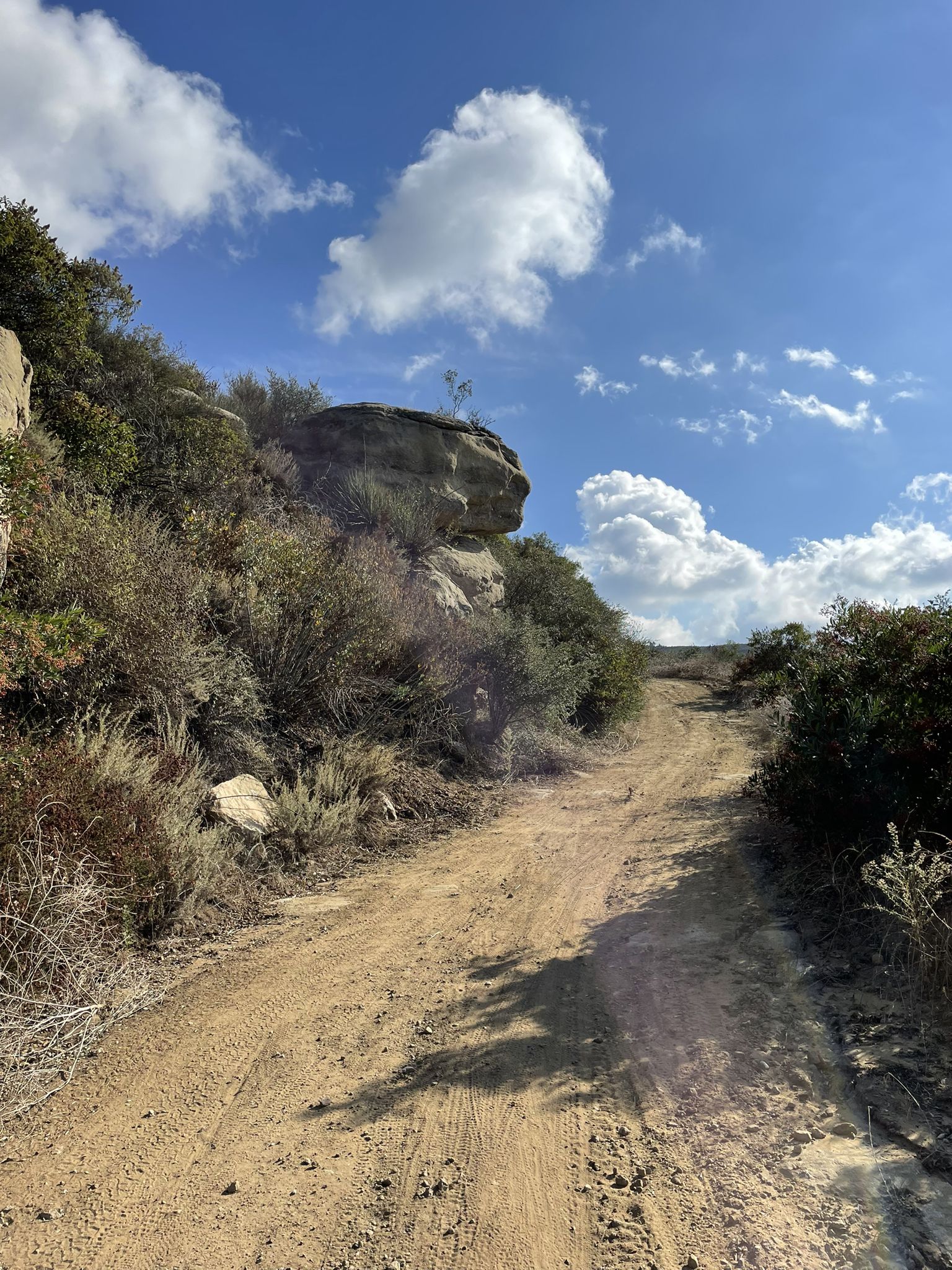 Dirt road ascending a rocky, brush-covered hillside under a bright, cloudy sky. - Sierra Peak via Coal Canyon Trail Loop