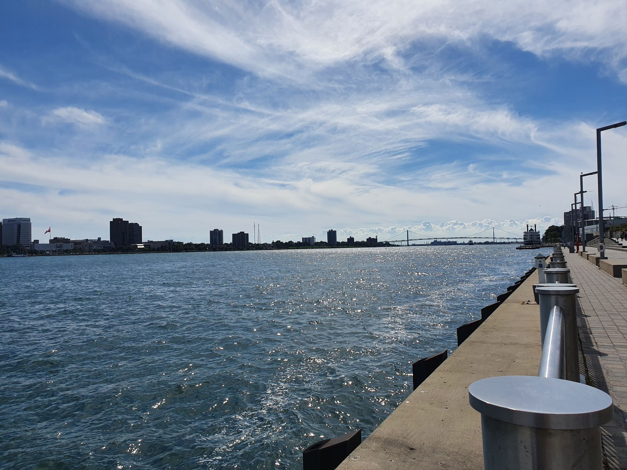 Concrete pier alongside a wide river with a distant city and bridge. - Detroit Riverwalk Viewpoint – William G. Milliken State Park loop from Detroit