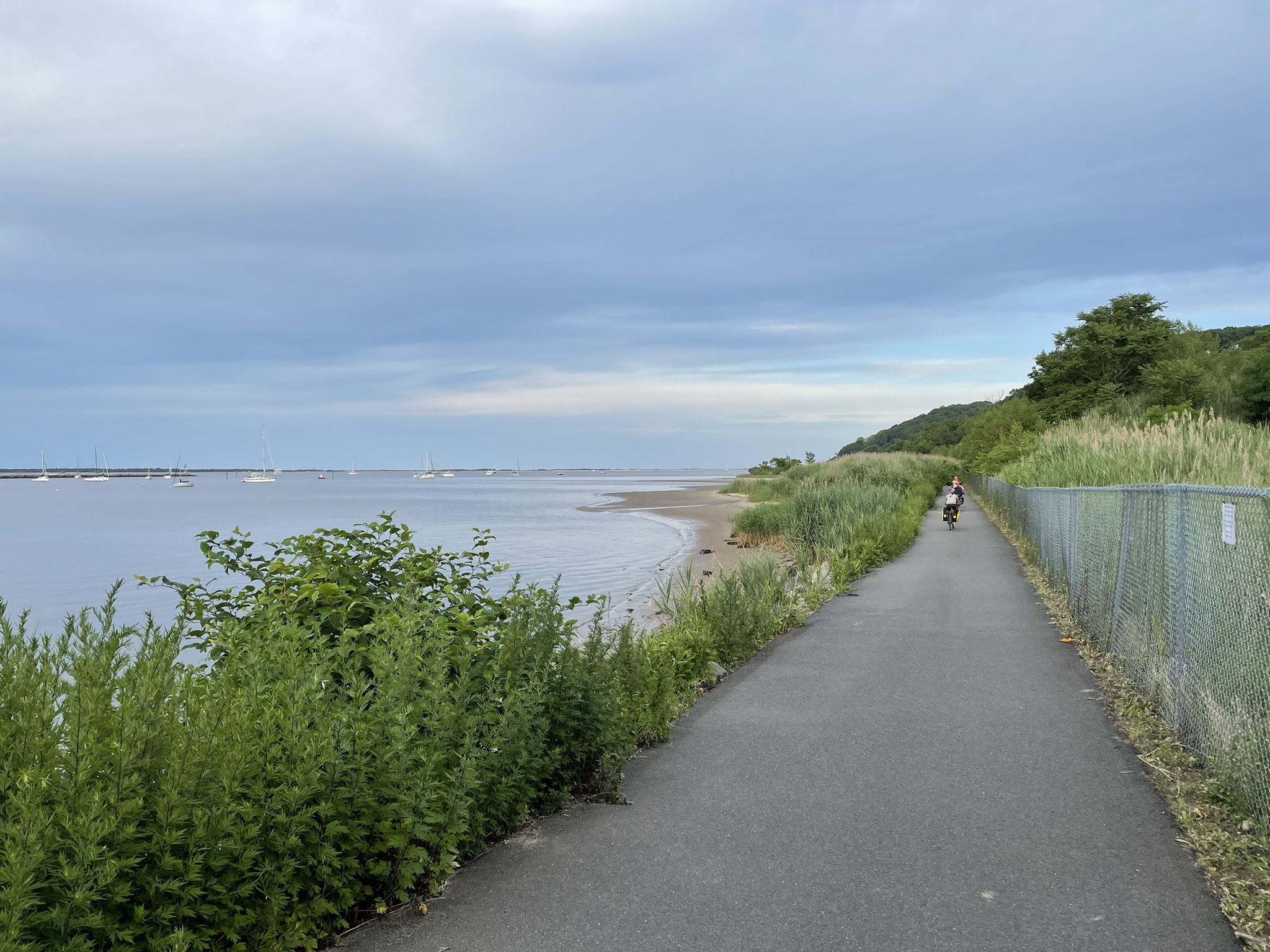 Paved path alongside a bay with boats, green foliage, and a person walking. - Henry Hudson Trail Loop