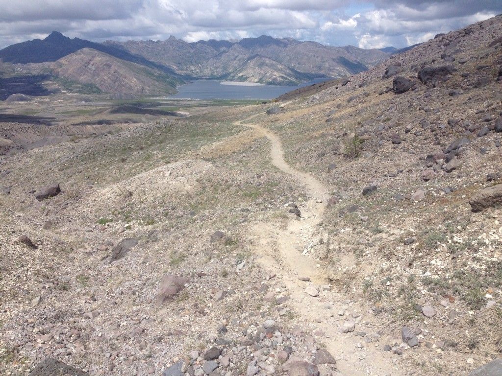 A winding dirt path through a dry, rocky landscape towards a lake. - Loowit Trail