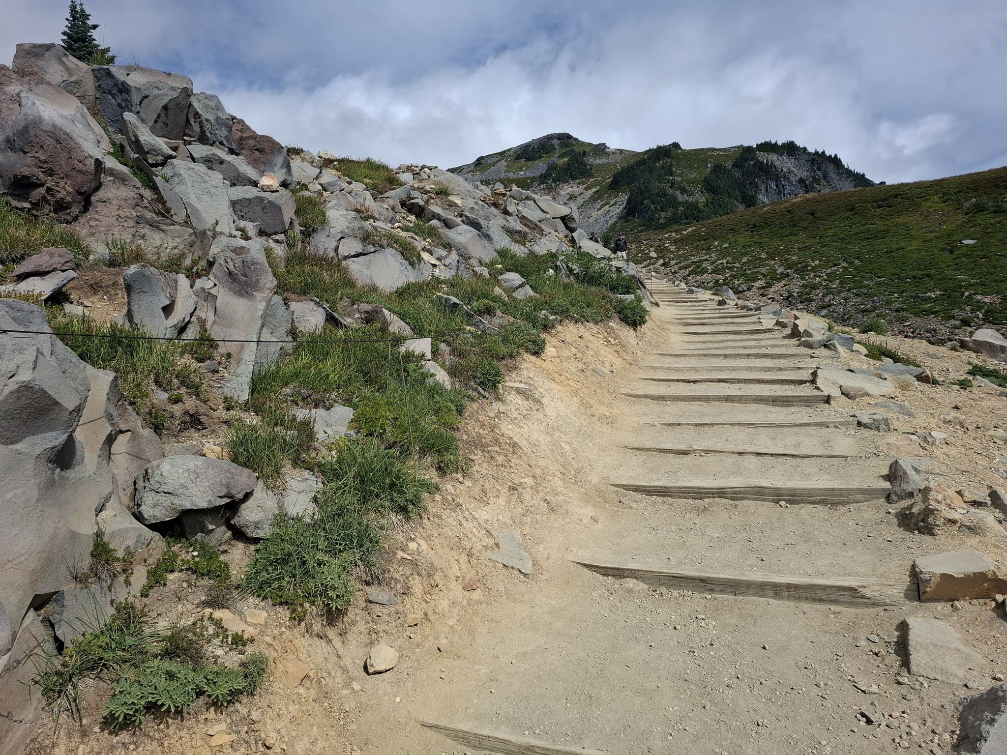 A dirt path with wooden steps ascends a rocky, grassy hillside under a cloudy sky. - Panorama Point via Skyline Trail