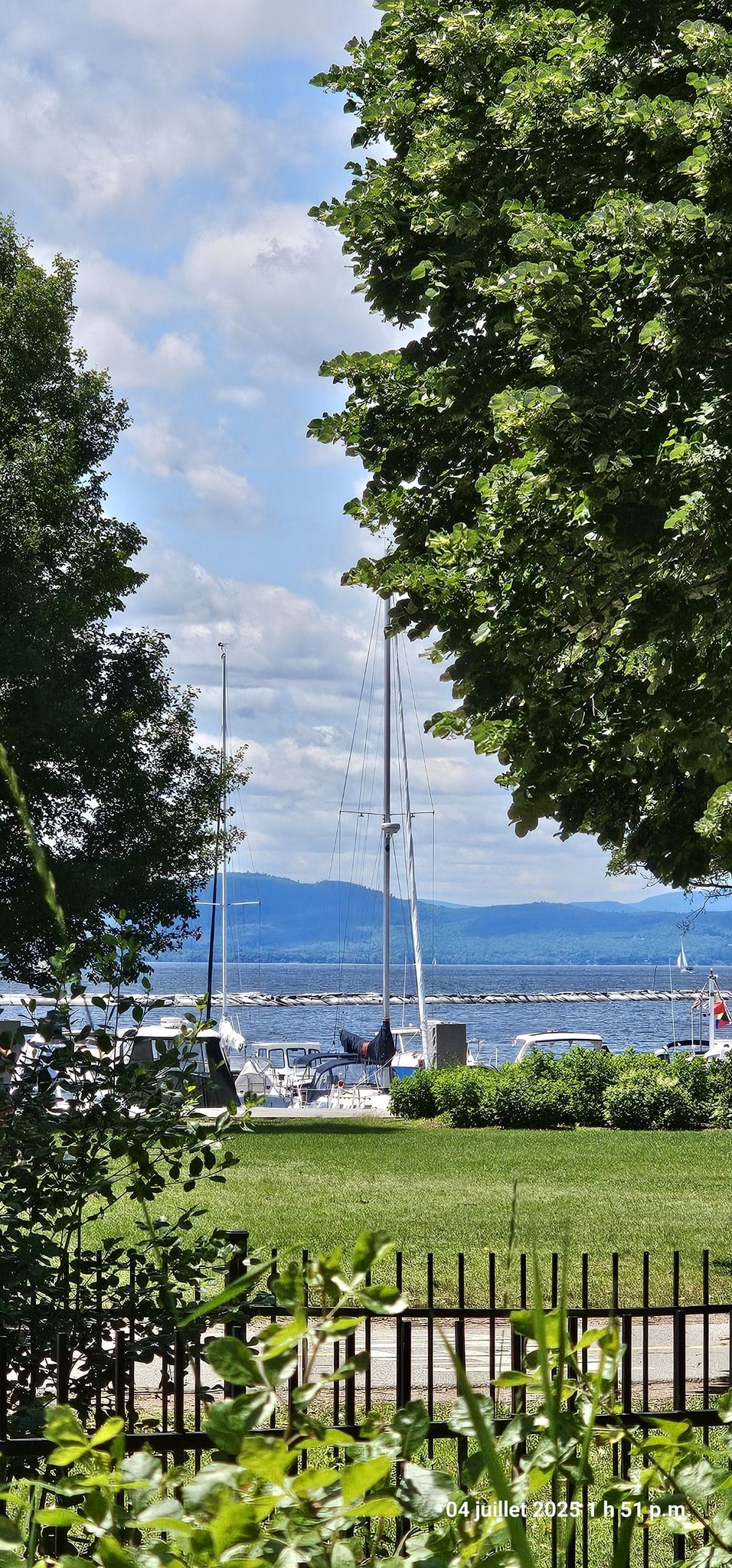 Sailboats are docked in a marina, visible through green trees, with distant mountains. - Waterfront Park – Granite Steps loop from South Burlington