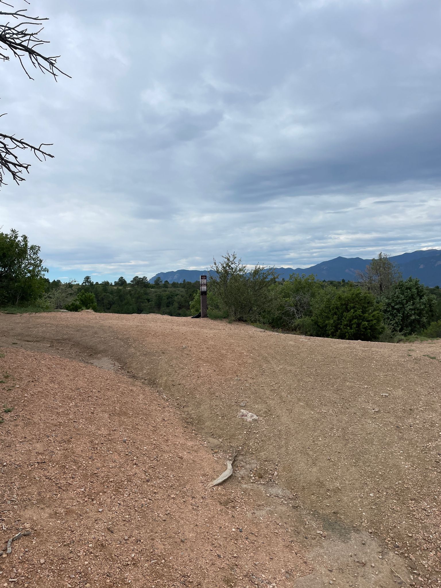 A dirt path leads to a trail sign, with mountains and trees in the distance. - Outer Loop