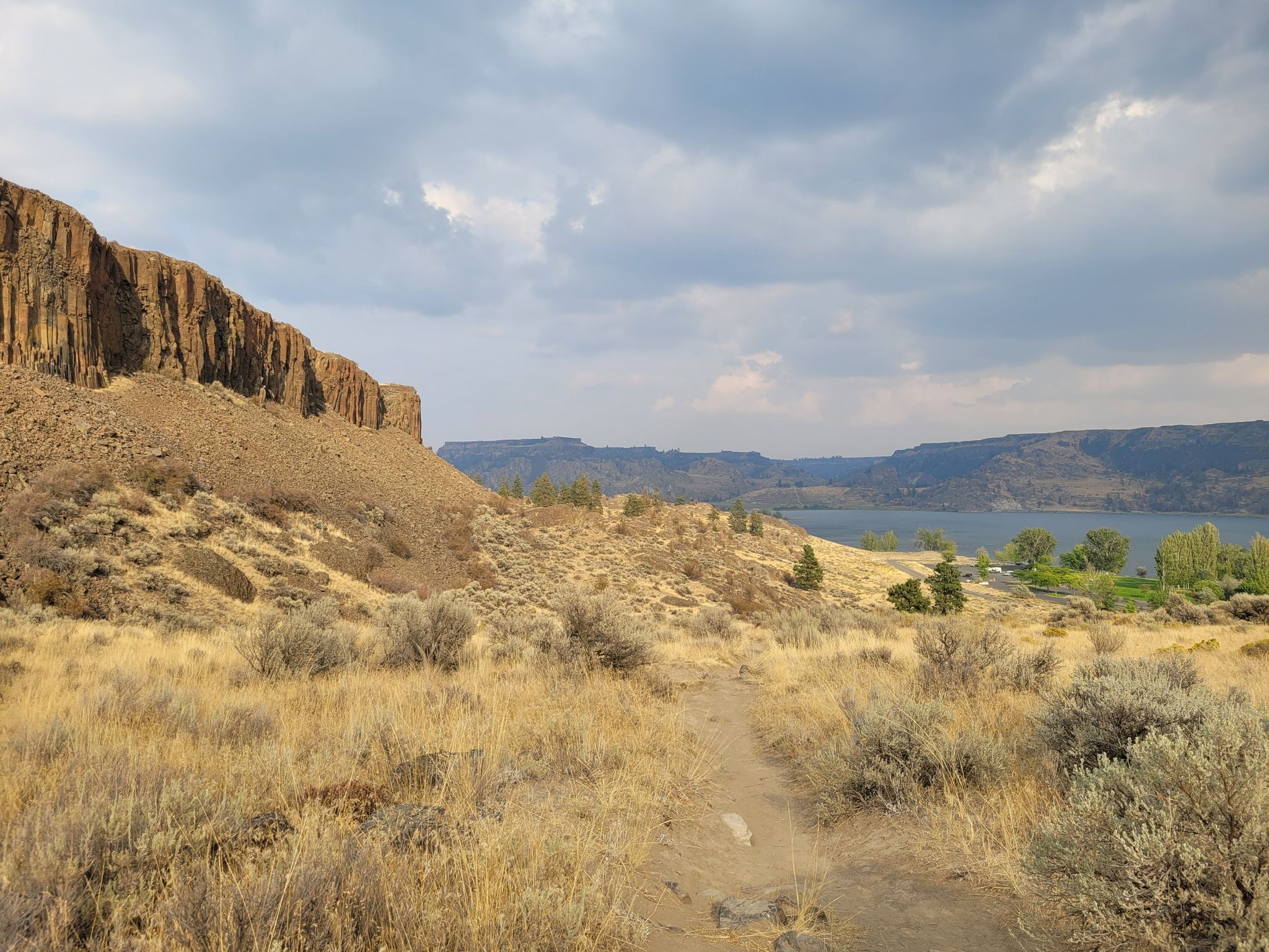 A dirt path winds through dry grass and shrubs towards a lake and cliffs. - Steamboat Rock Trail