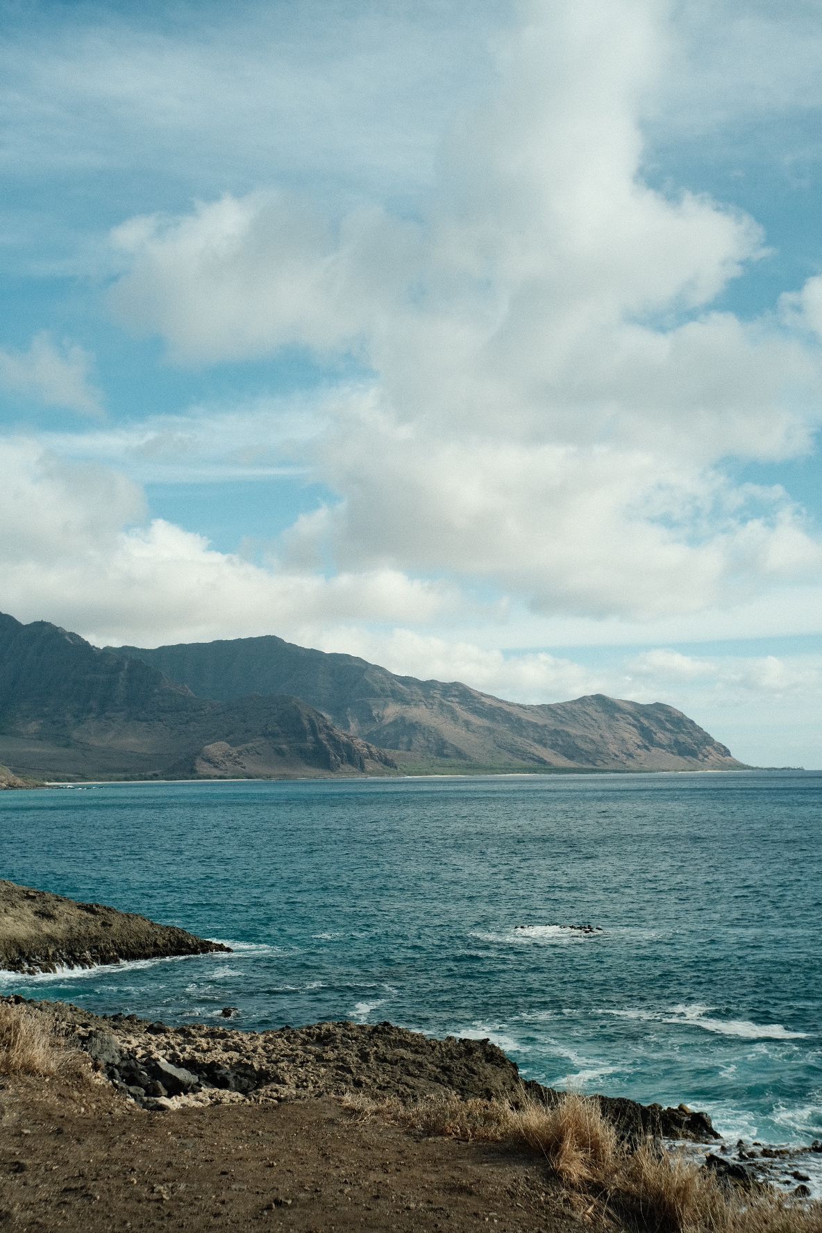 Blue ocean meets rugged mountains under a partly cloudy sky. - Nānākuli loop