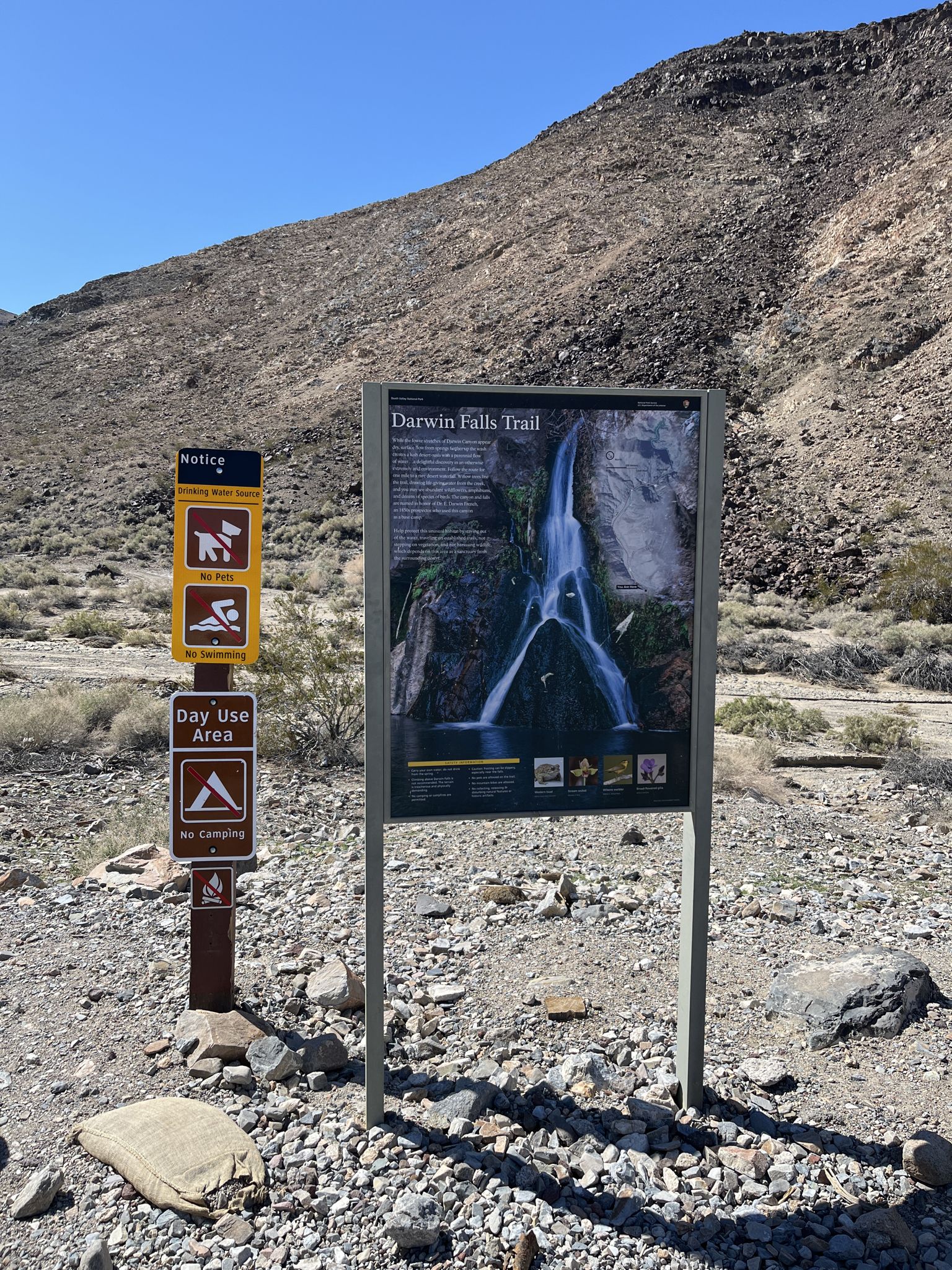 An information sign and trail markers stand in a rocky desert landscape. - Upper Darwin Falls via Old Toll Road