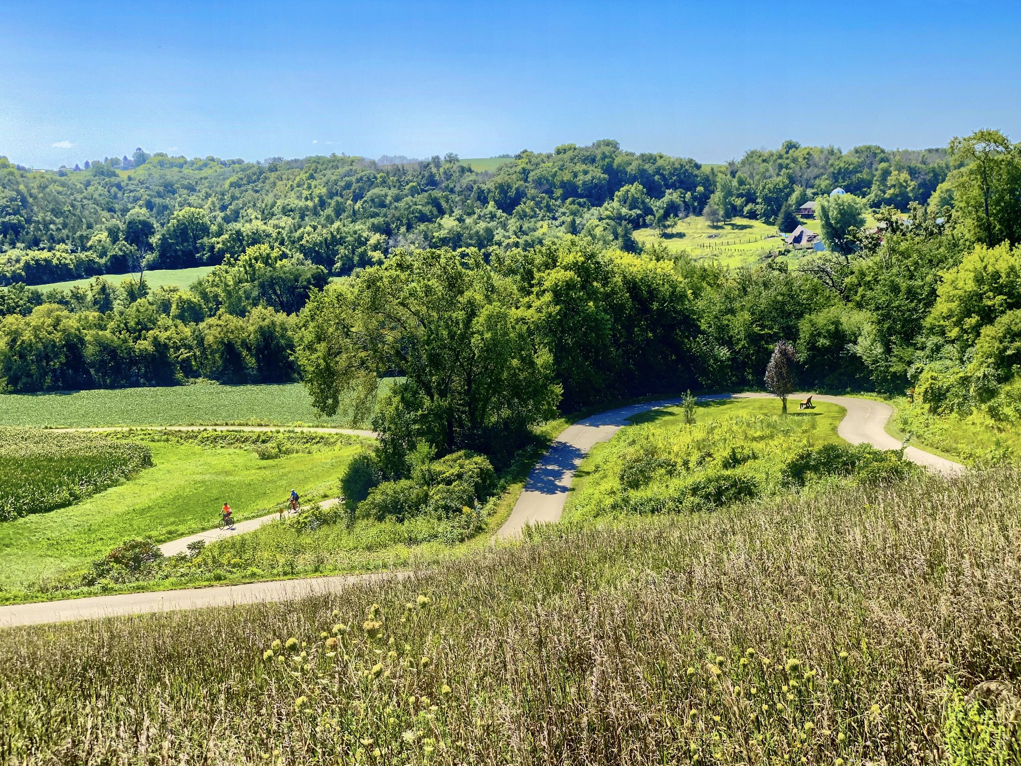 Elevated view of winding path through green fields, two cyclists, sunny. - Trout Run Trail
