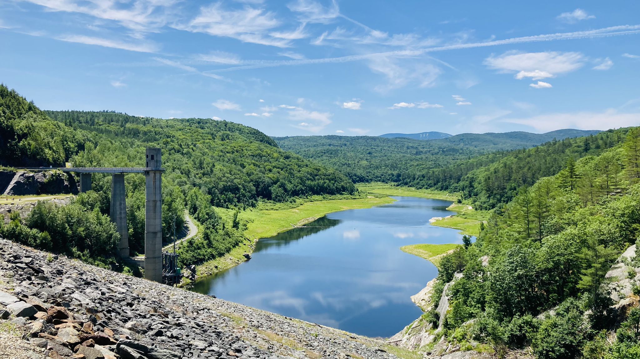 A reservoir with a concrete structure, surrounded by green forested hills under a blue sky. - West River Trail