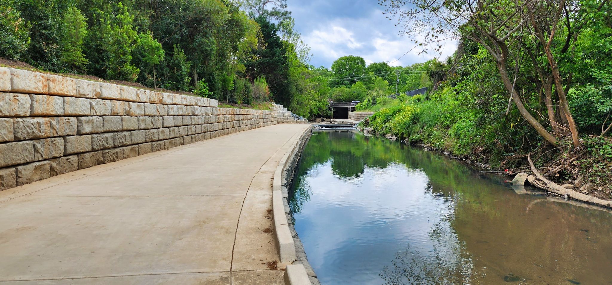 Paved path alongside a stone wall and calm river under cloudy skies. - Little Sugar Creek Greenway – Alexander Street Park loop from Arrowood