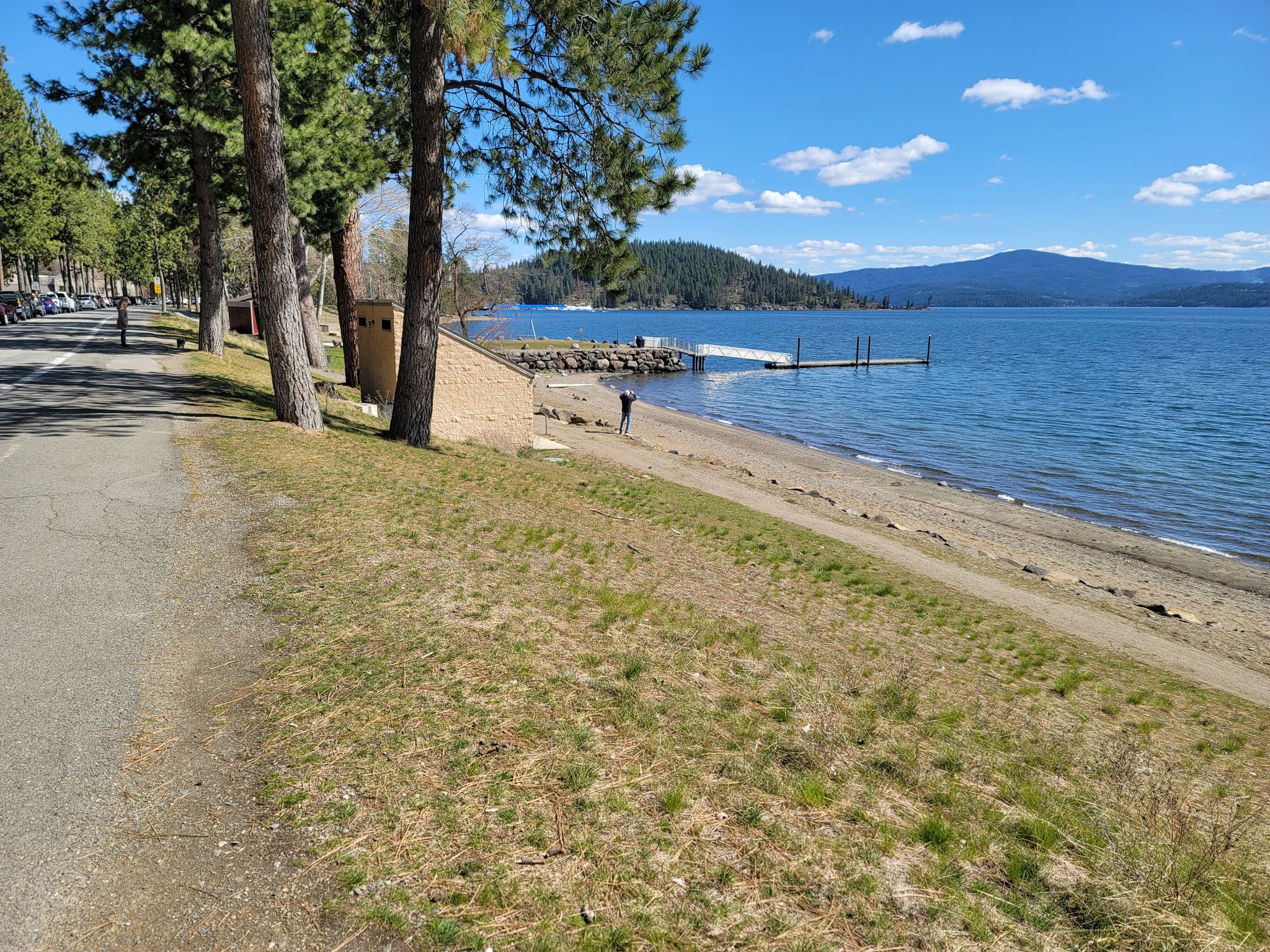 Paved path beside a sandy lake beach with one person and forested hills. - North Idaho Centennial Trail