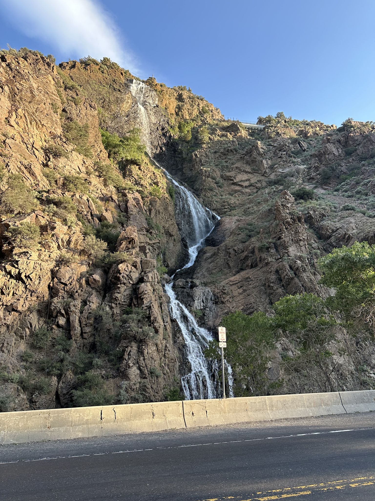 Waterfall cascades down a rocky mountain beside a road under a bright sky. - Denver and Rio Grande Western Rail Trail Loop