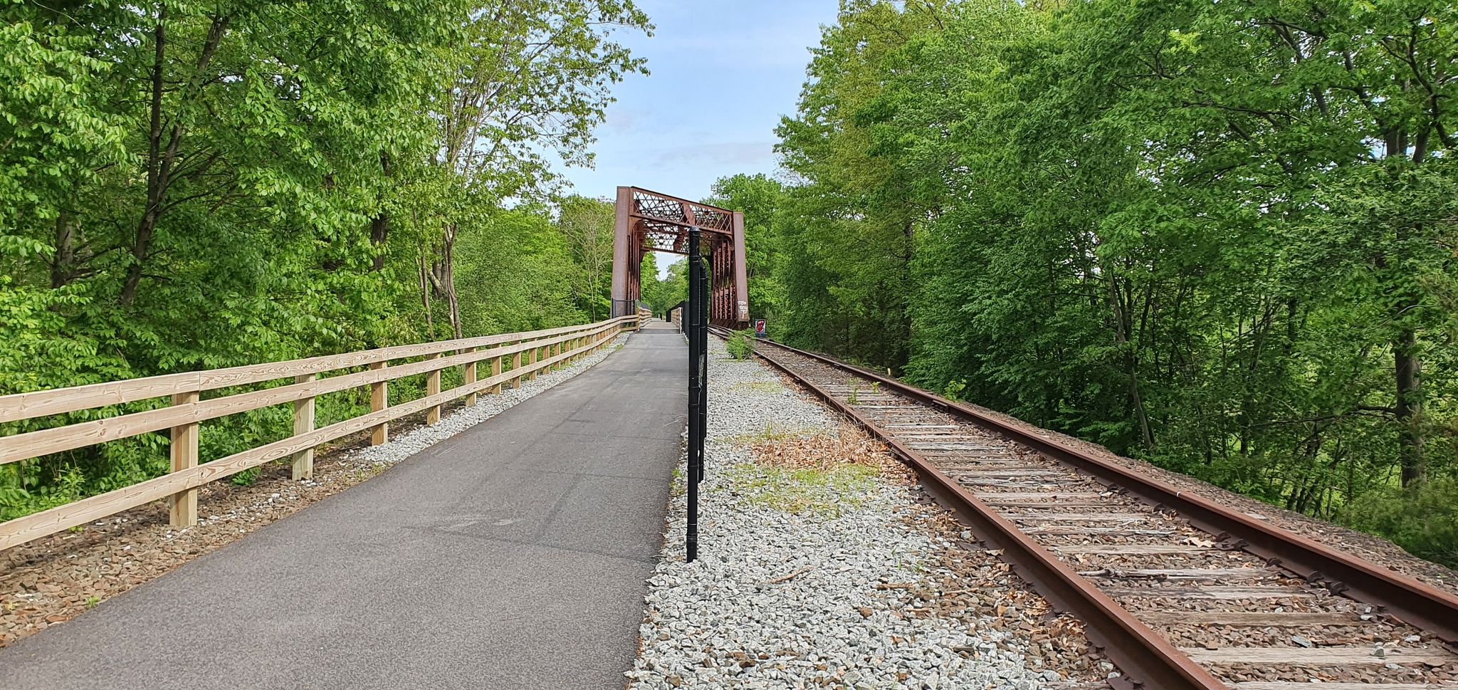 Paved path and railroad tracks lead to a metal bridge through green trees. - Stewart's Farm loop from Mahopac
