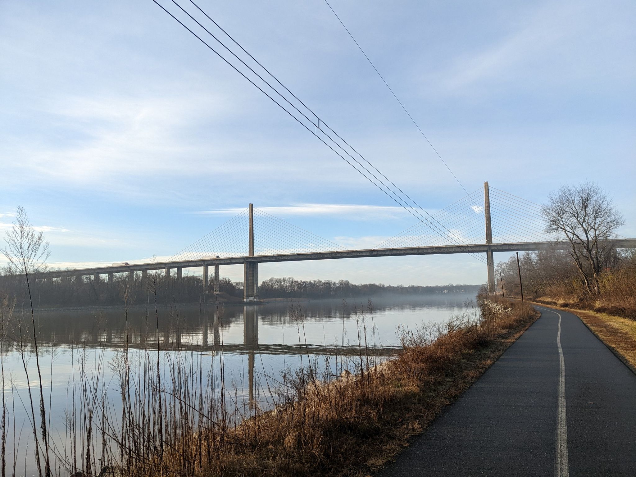 A paved path next to a calm river with a distant cable-stayed bridge. - Summit Pointe – Mike Castle Trail loop from North Saint Georges
