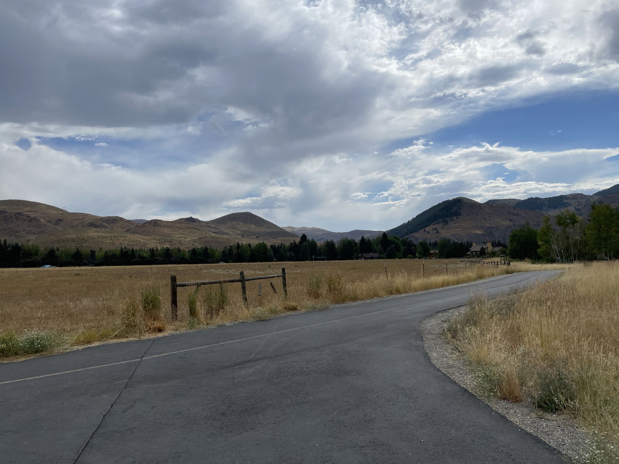 Paved road curves through dry fields towards distant mountains under a cloudy sky. - Penny Mountain – Big Wood River loop from Sun Valley