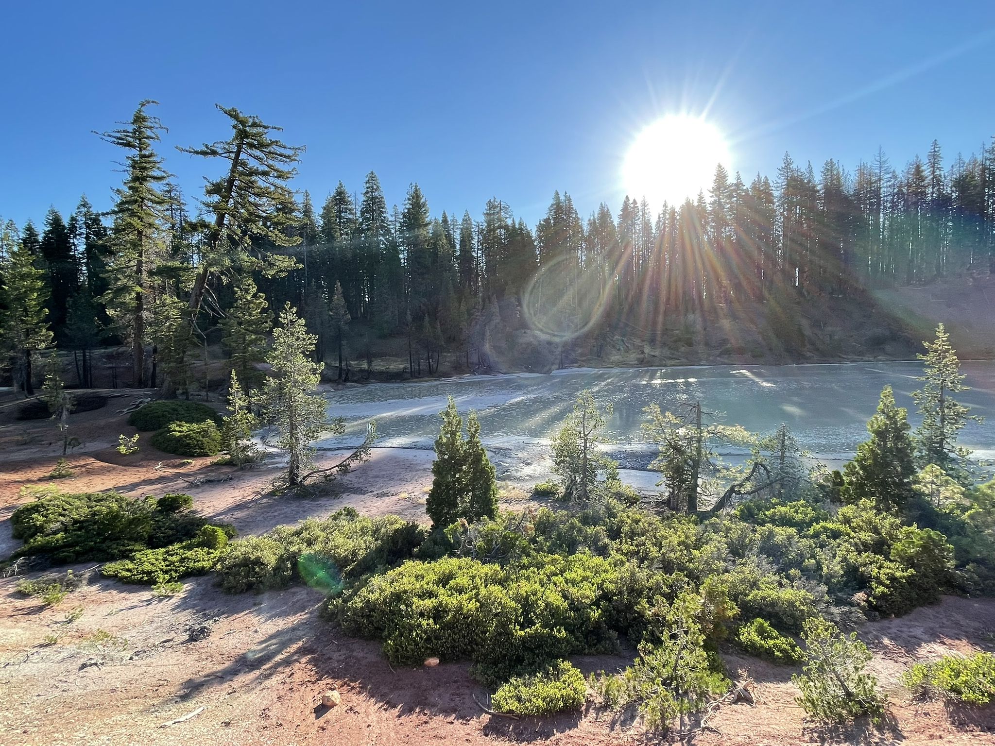 Bright sunburst over a steaming lake surrounded by green forest and reddish ground. - Boiling Springs Lake Trail