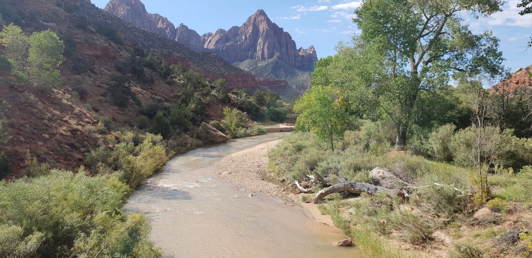 A river flows through a green valley with rugged mountains under a blue sky. - Hurricane, Utah to Zion National Park