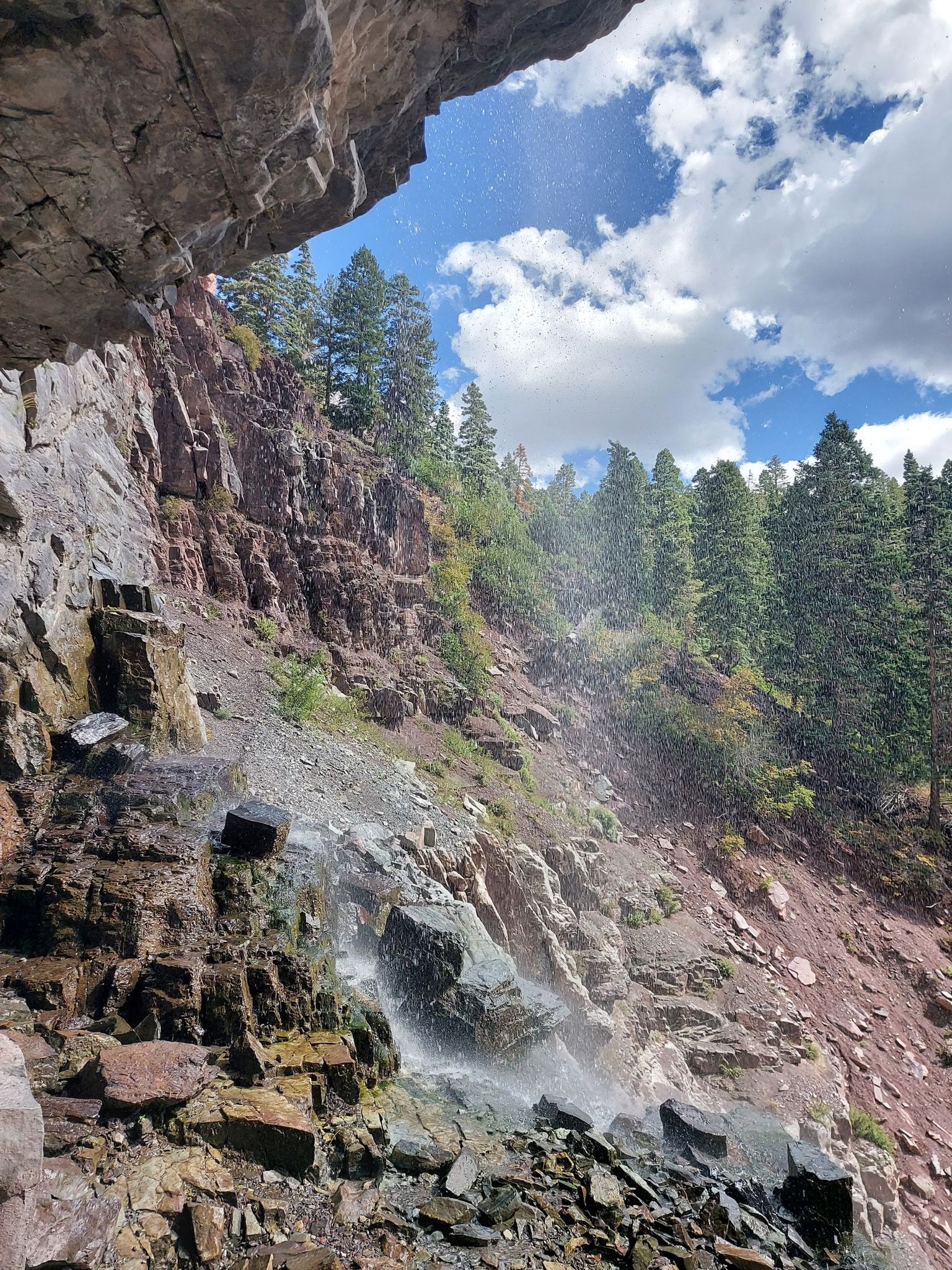 Waterfall misting over a rocky cliff with green trees under a cloudy sky. - Lower Cascade Falls Trail