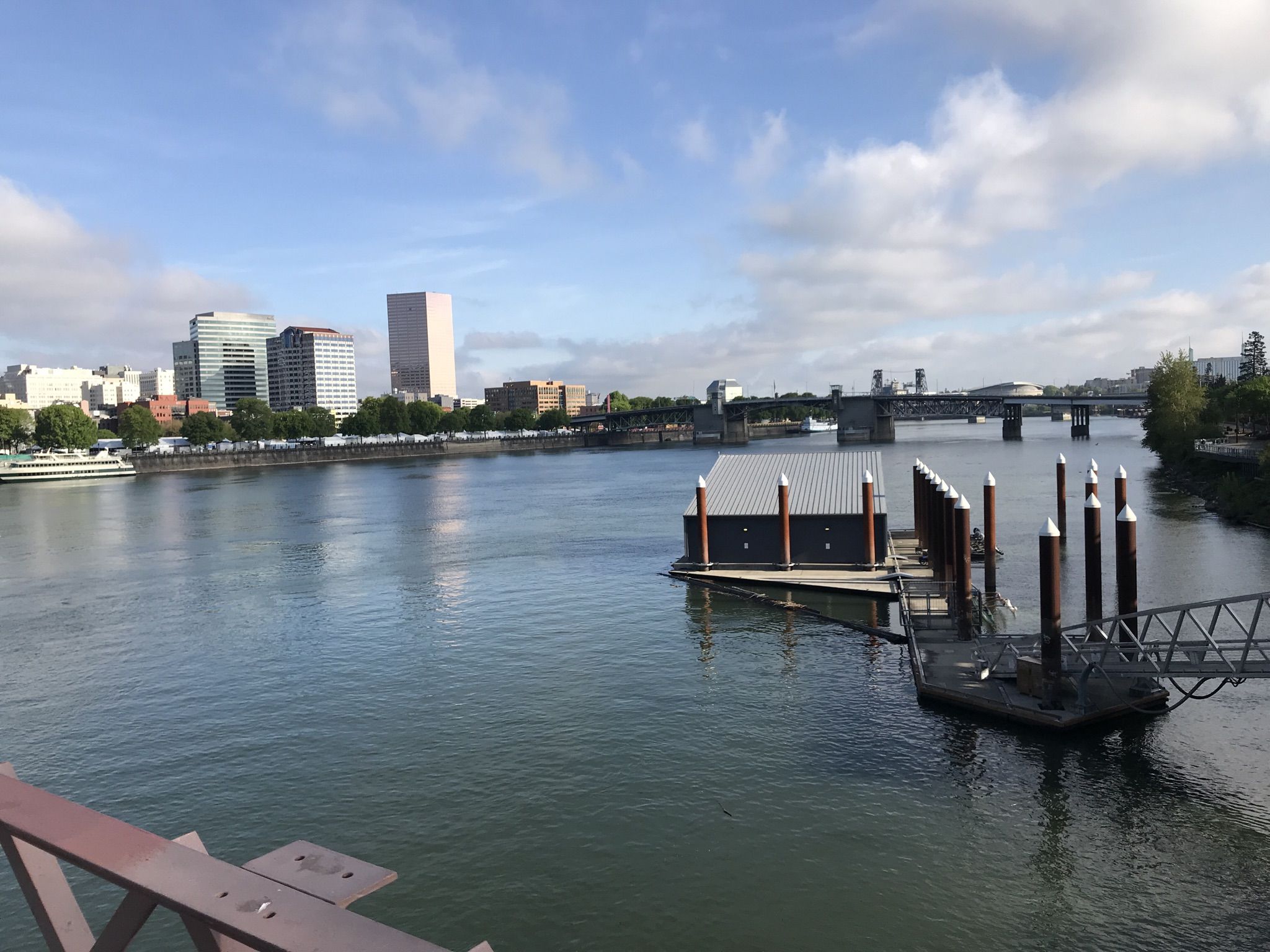 A city skyline overlooks a river with a floating dock on a bright day. - View of Saint Johns Bridge – Hawthorne Bridge loop from Goose Hollow/Southwest Jefferson Street