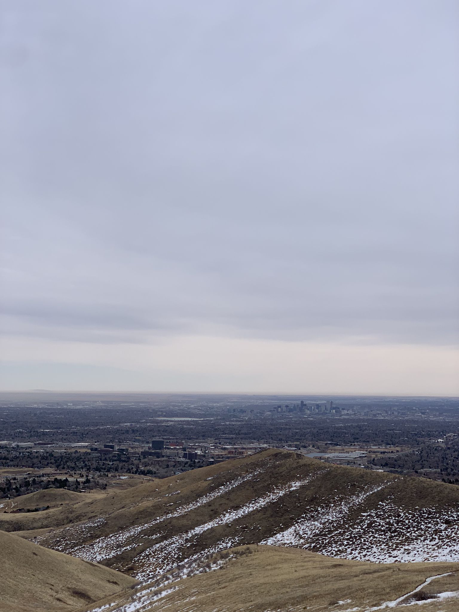 Panoramic view of a distant city beyond snow-dusted hills under grey sky. - North Trail loop from West Pleasant View