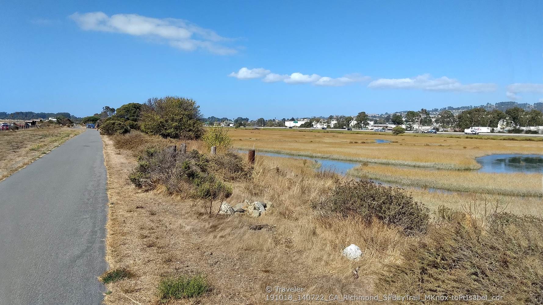Paved path alongside a marshy wetland with distant buildings under a blue sky. - Aquatic Park North Trailhead – Point Isabel loop from Berkeley
