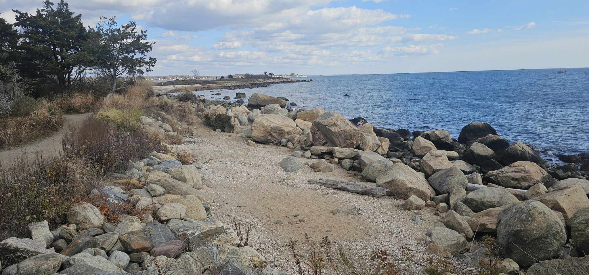 Rocky shoreline with a sandy path next to calm blue ocean under a cloudy sky. - Hammonasset Point via Hammonasset Beach loop