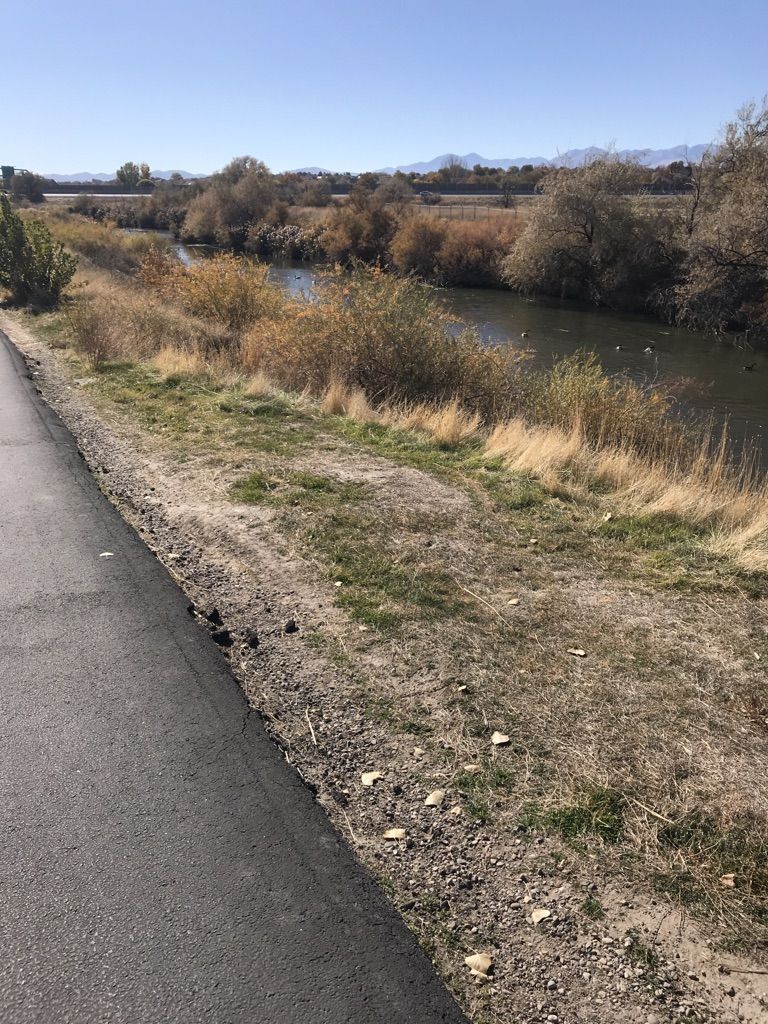 Sunny view of a paved path beside a river with dry brush and distant mountains. - Jordan River Parkway – View of the Jordan River loop from Meadowbrook