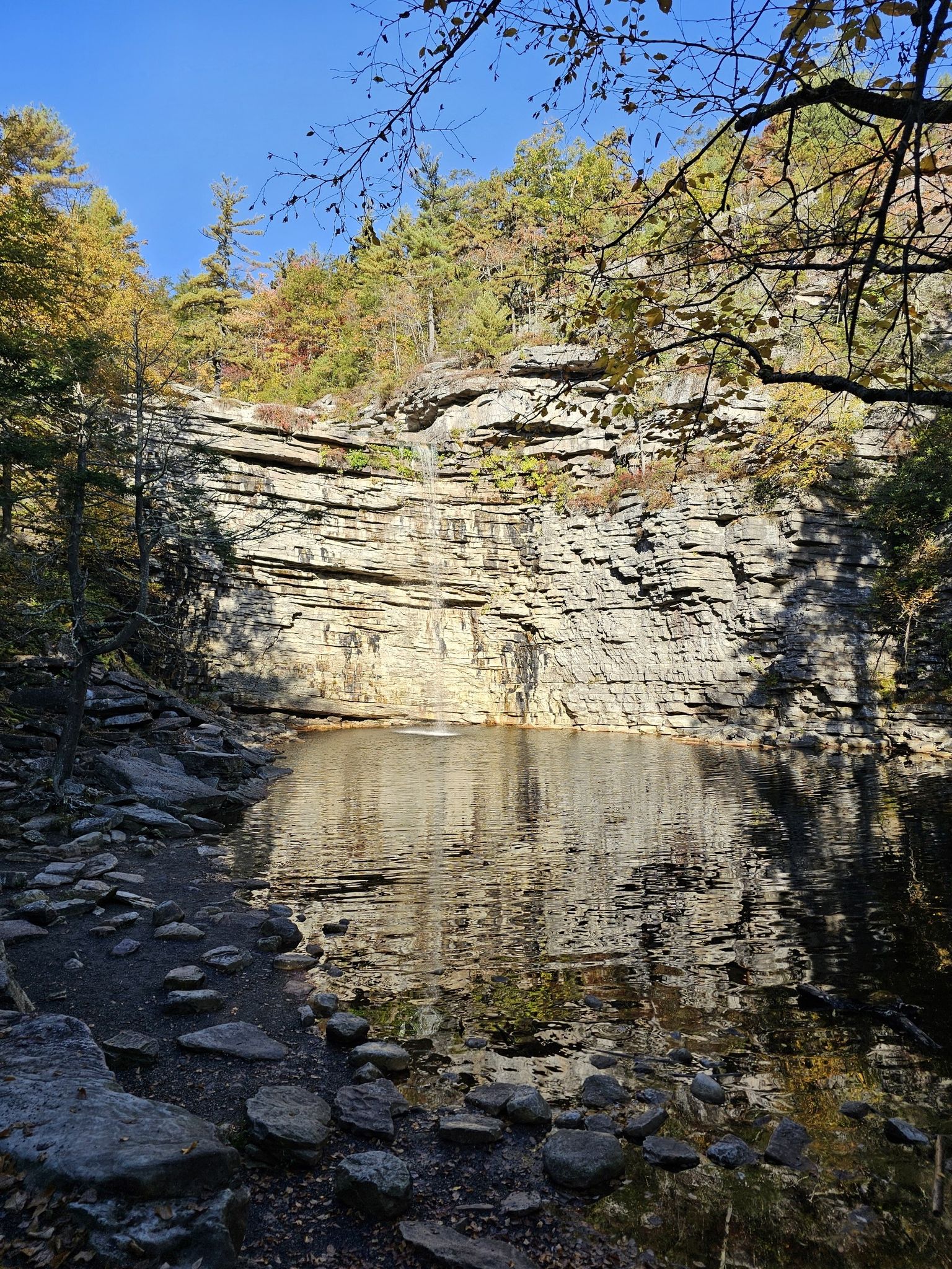 Calm pool at the base of a rocky cliff, surrounded by autumn trees under clear sky. - Awosting Falls loop from New Paltz