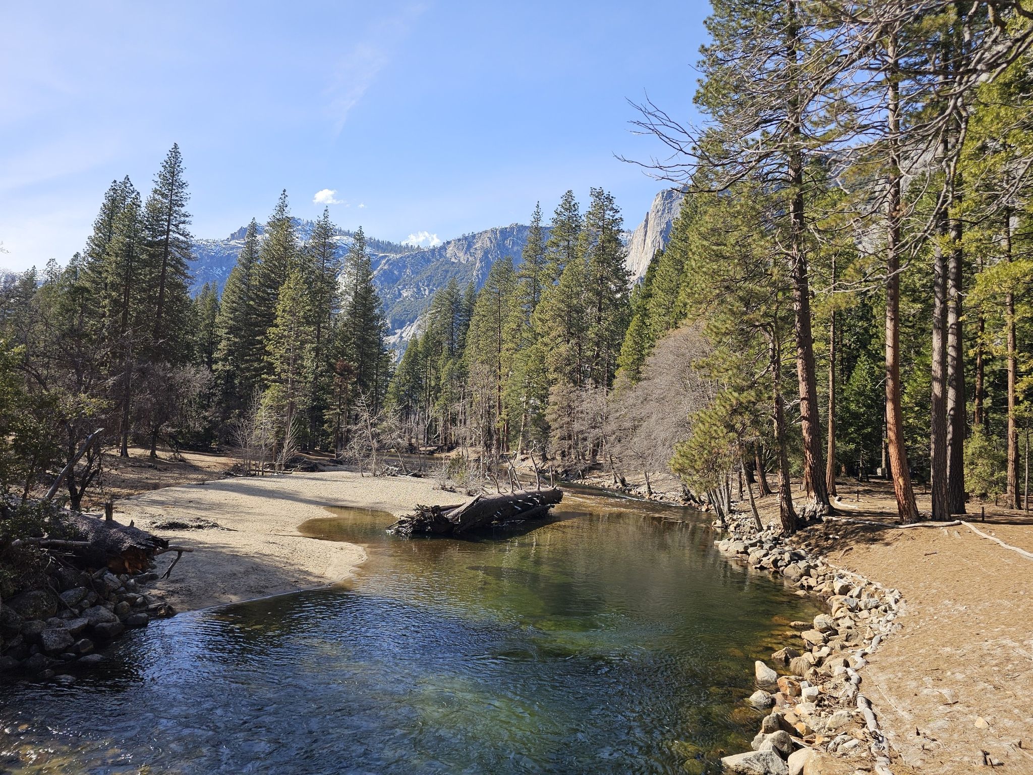 Clear river flowing through a forest with distant snow-capped mountains under bright sun. - Yosemite Valley Loop Trail