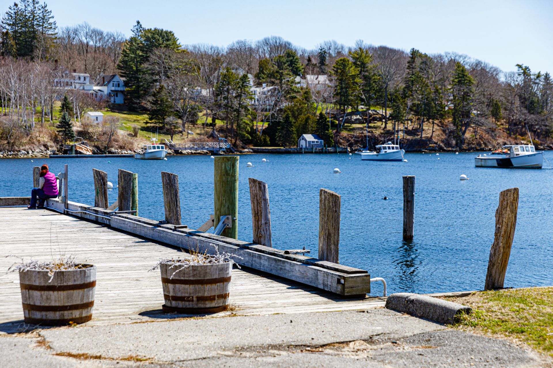 Person sits on a sunny wooden dock overlooking calm blue water and distant shore. - Rockport Harbor loop from Rockport