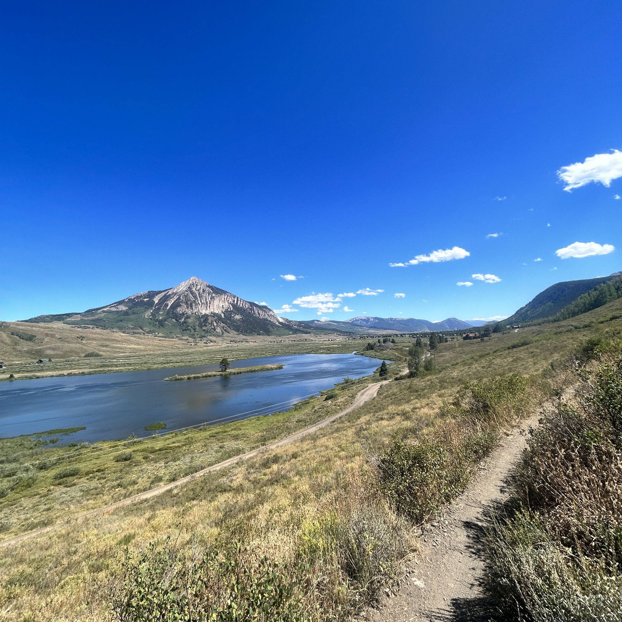 Dirt trail overlooks a serene lake and majestic mountain under a clear blue sky. - Snodgrass, Lupine, Lower Loop