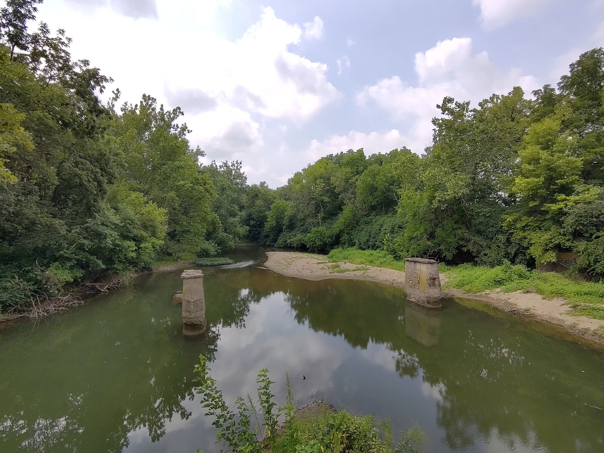 Calm river with concrete structures, lush green banks, under a cloudy sky. - Creekside Trail – Xenia Station loop from Southern Ohio Brewing
