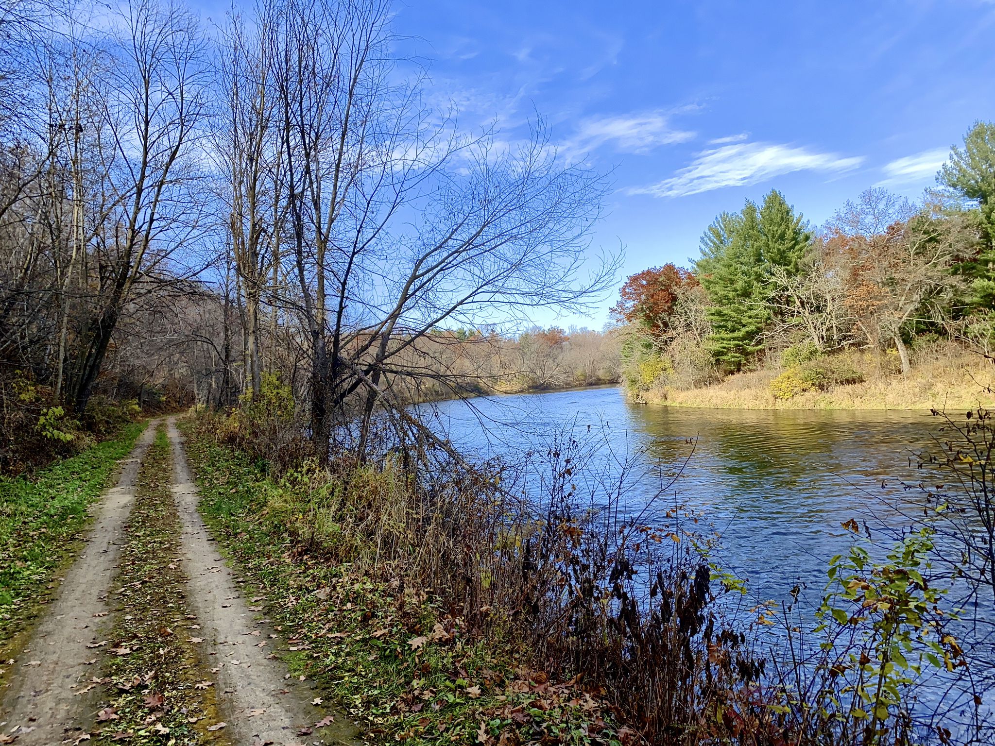 Dirt path beside a blue river, bare trees, and autumn foliage under clear sky. - Devil's Punchbowl loop from Menomonie
