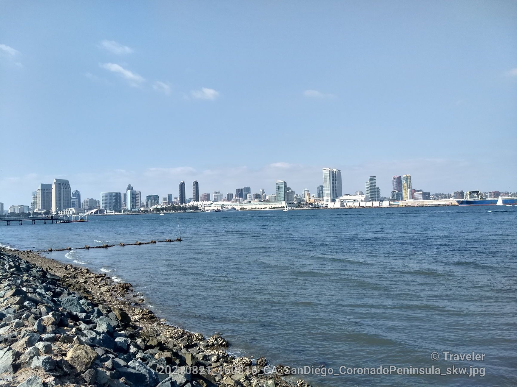 Distant city skyline across a wide body of water with a rocky shore. - Coronado Ferry Landing – View of Downtown San Diego loop from Coronado