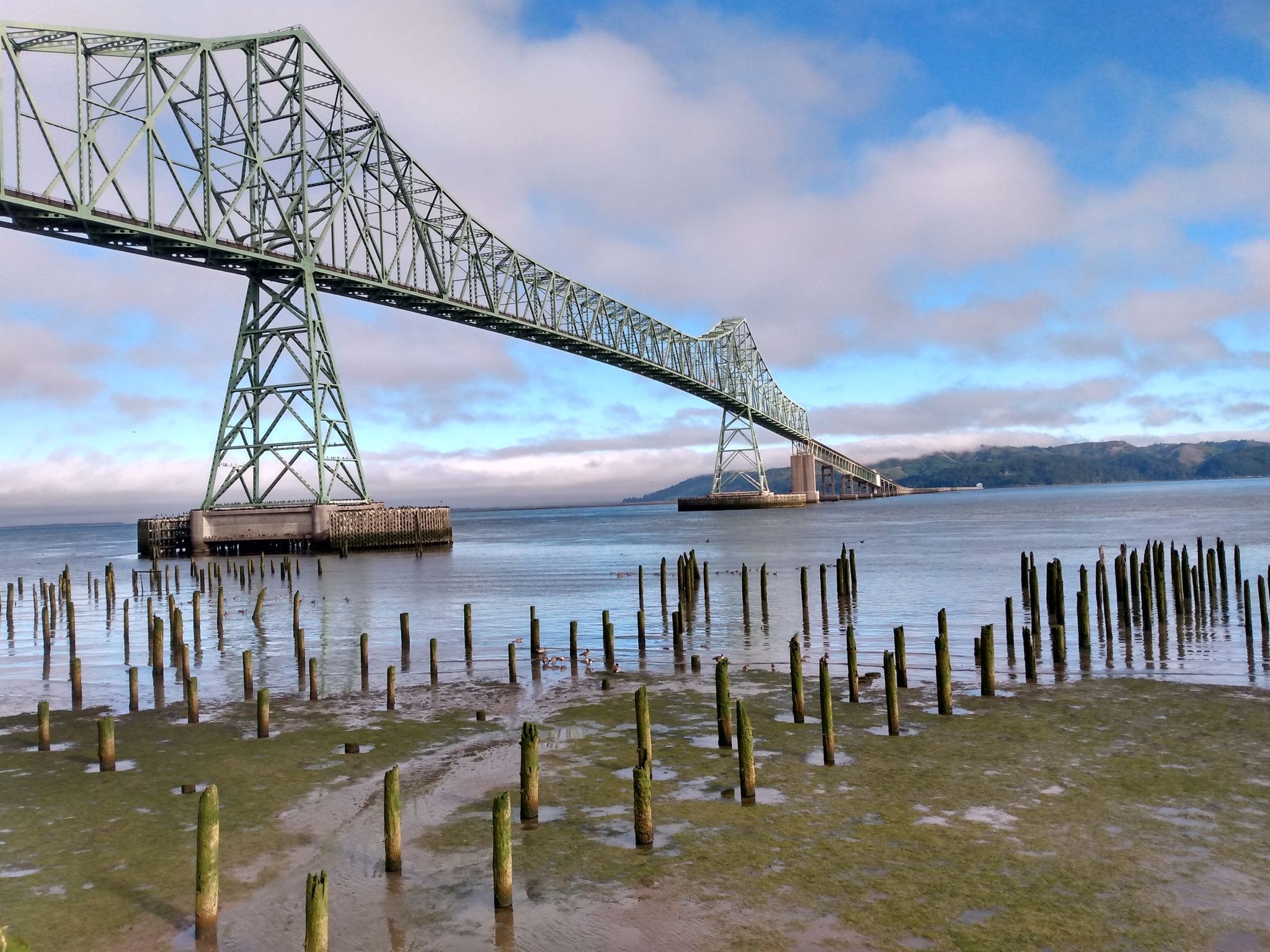 Green steel bridge over water, many old pilings, partly cloudy sky. - Astoria Riverwalk Trail