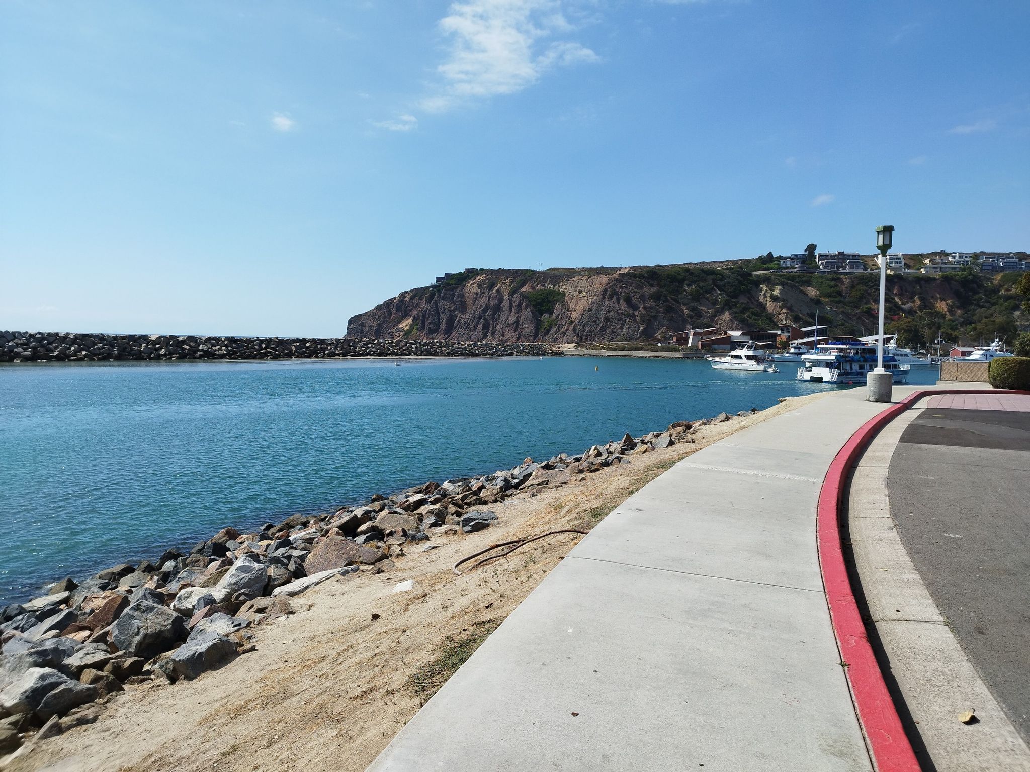 Sunny harbor with boats, a rocky shore, and a paved walkway. - Dana Point Harbor Loop