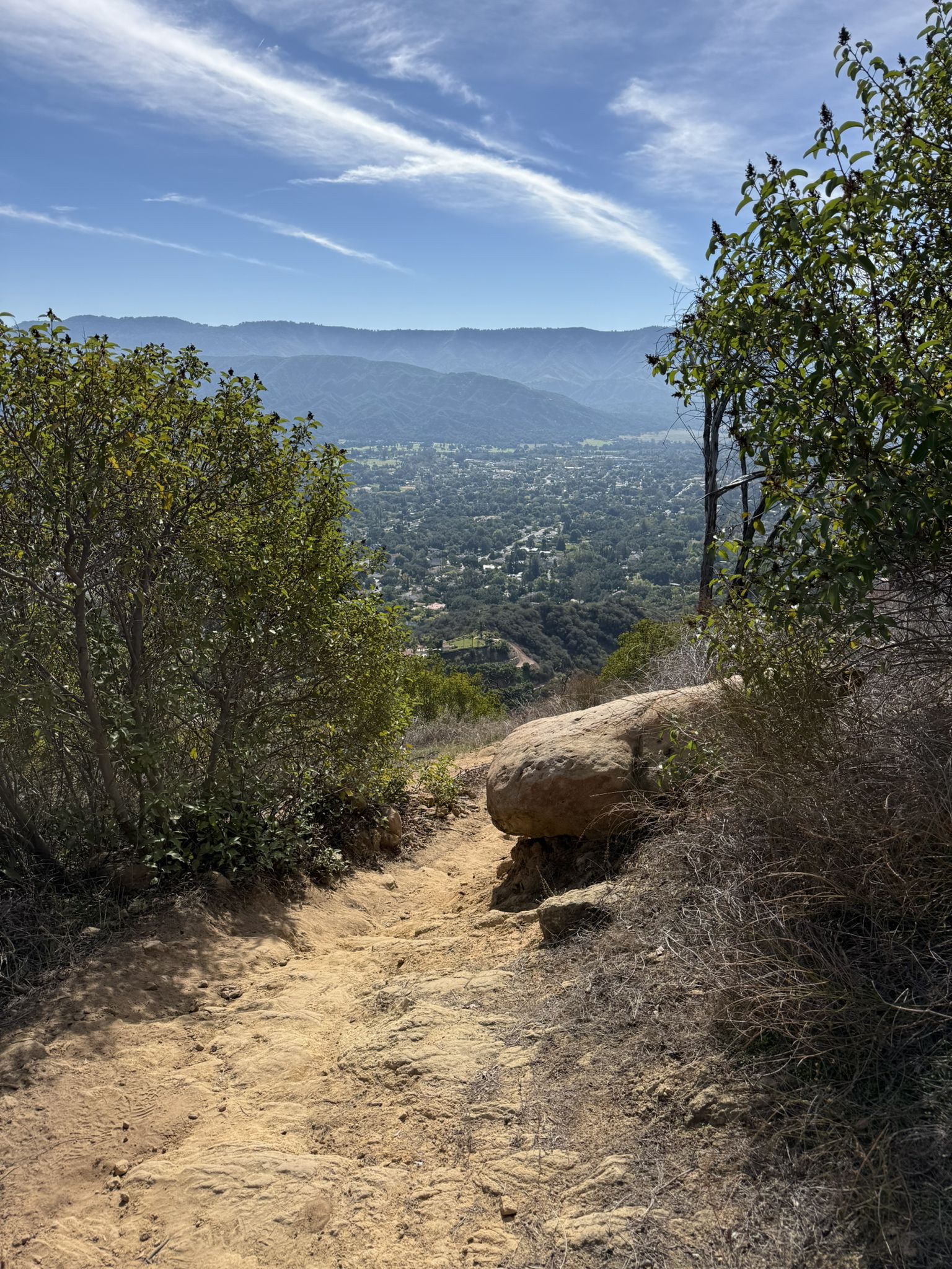 Dirt path descends into a sunny valley with distant mountains and town. - Pratt, Foothill, and Fox Canyon Trails Loop