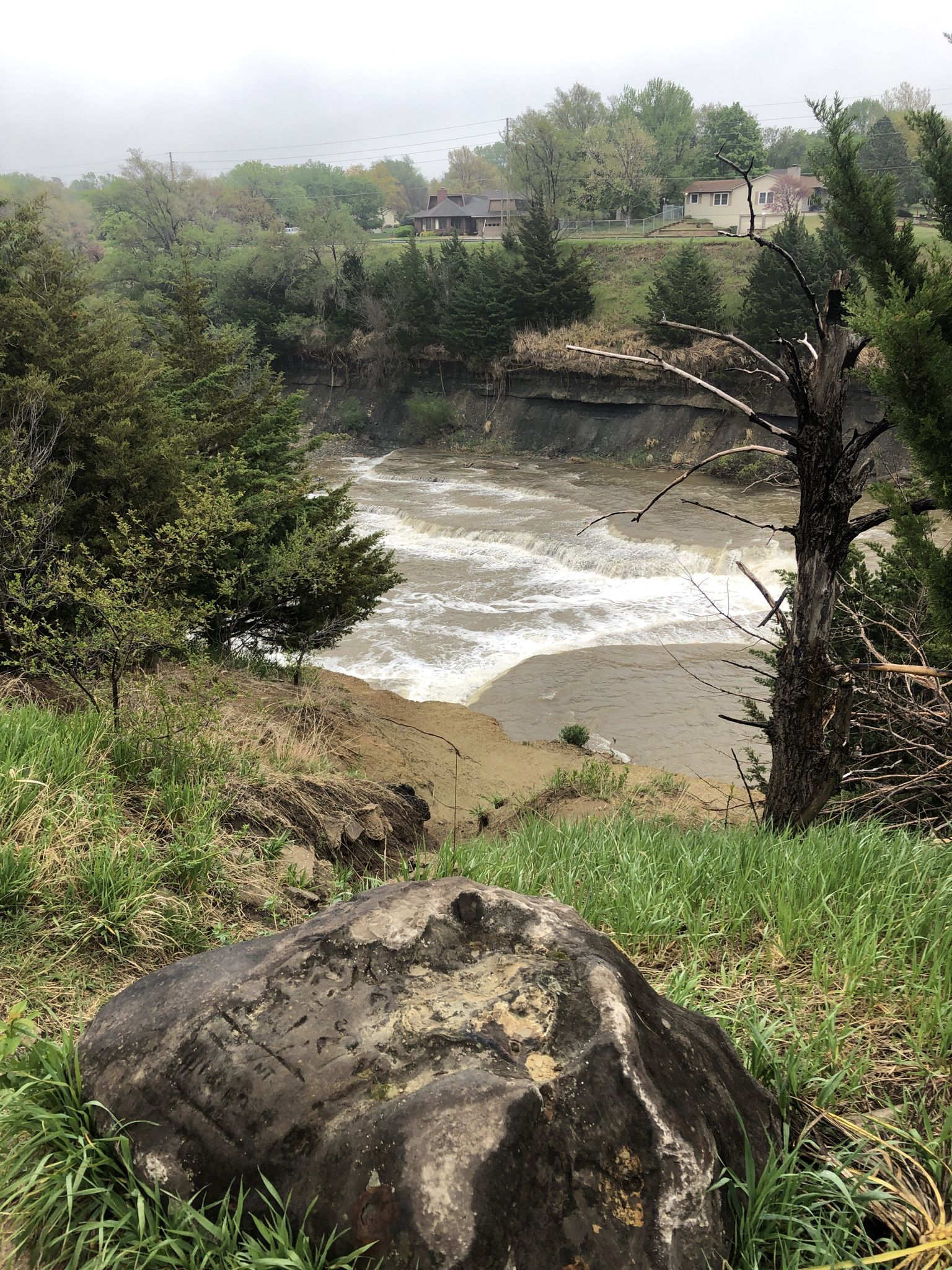Rushing river through a wooded landscape under an overcast sky, large boulder foreground. - Indian Rock Trail