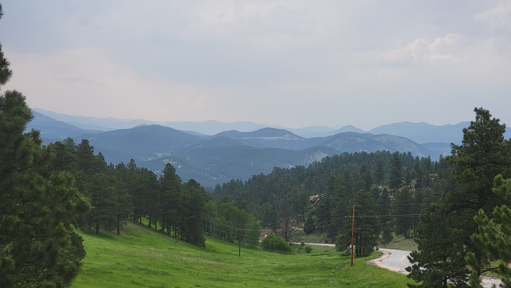 Green hillside and forested mountains under a cloudy sky, with a road. - Independence Mountain Trail