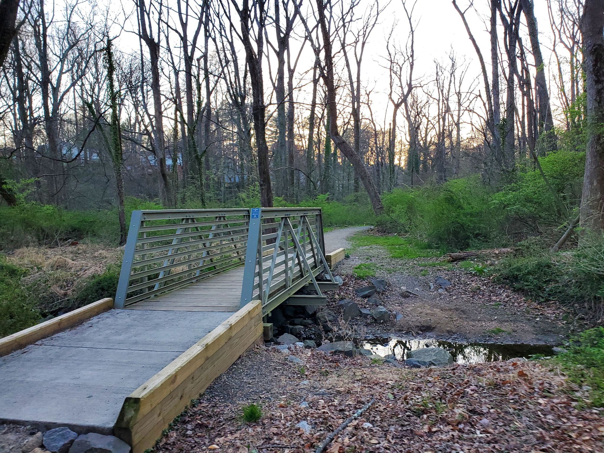 A metal and wood bridge crosses a small stream in a wooded landscape. - Holmes Run Trail and Roundtree Park Loop
