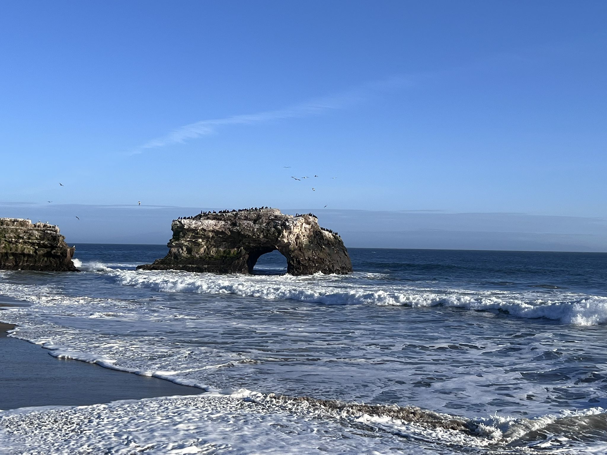 Ocean waves crash near a large rock arch under a clear blue sky. - Moore Creek Trail