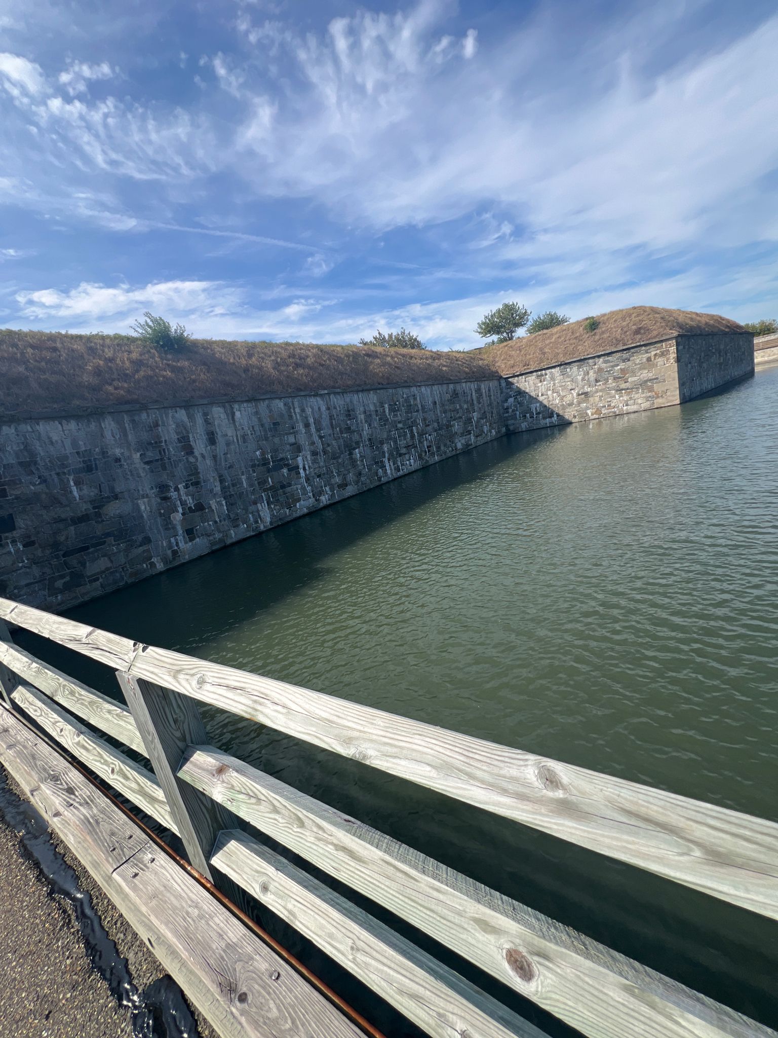 Sunny view of a stone wall and water moat from a wooden bridge. - Fort Monroe Seawall Trail