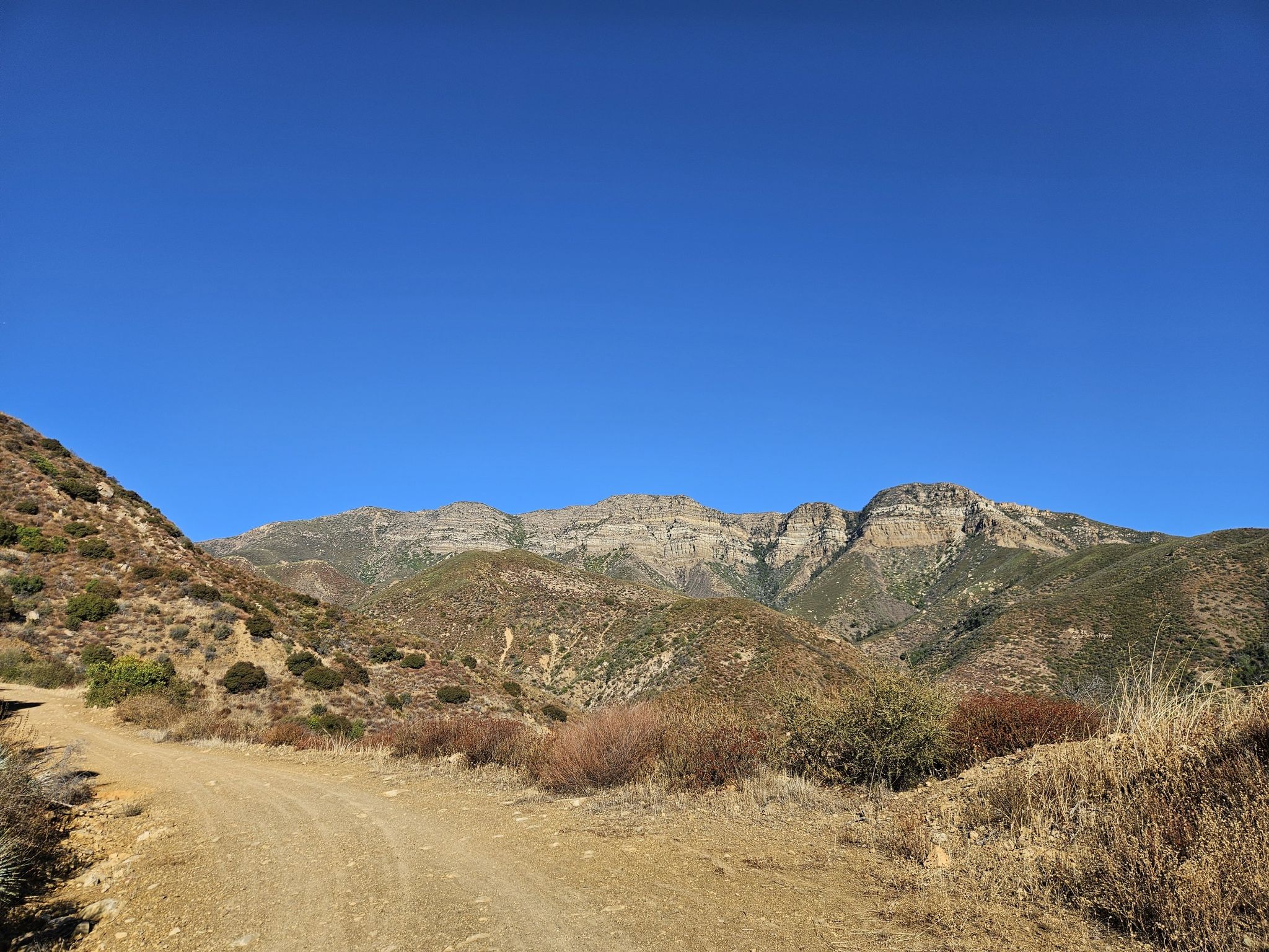 Wide dirt road through scrubby hills leading to rugged mountains under clear sky. - Sisar Canyon Road – Sisar Canyon Road loop from Stagecoach Station Upper Ojai Market