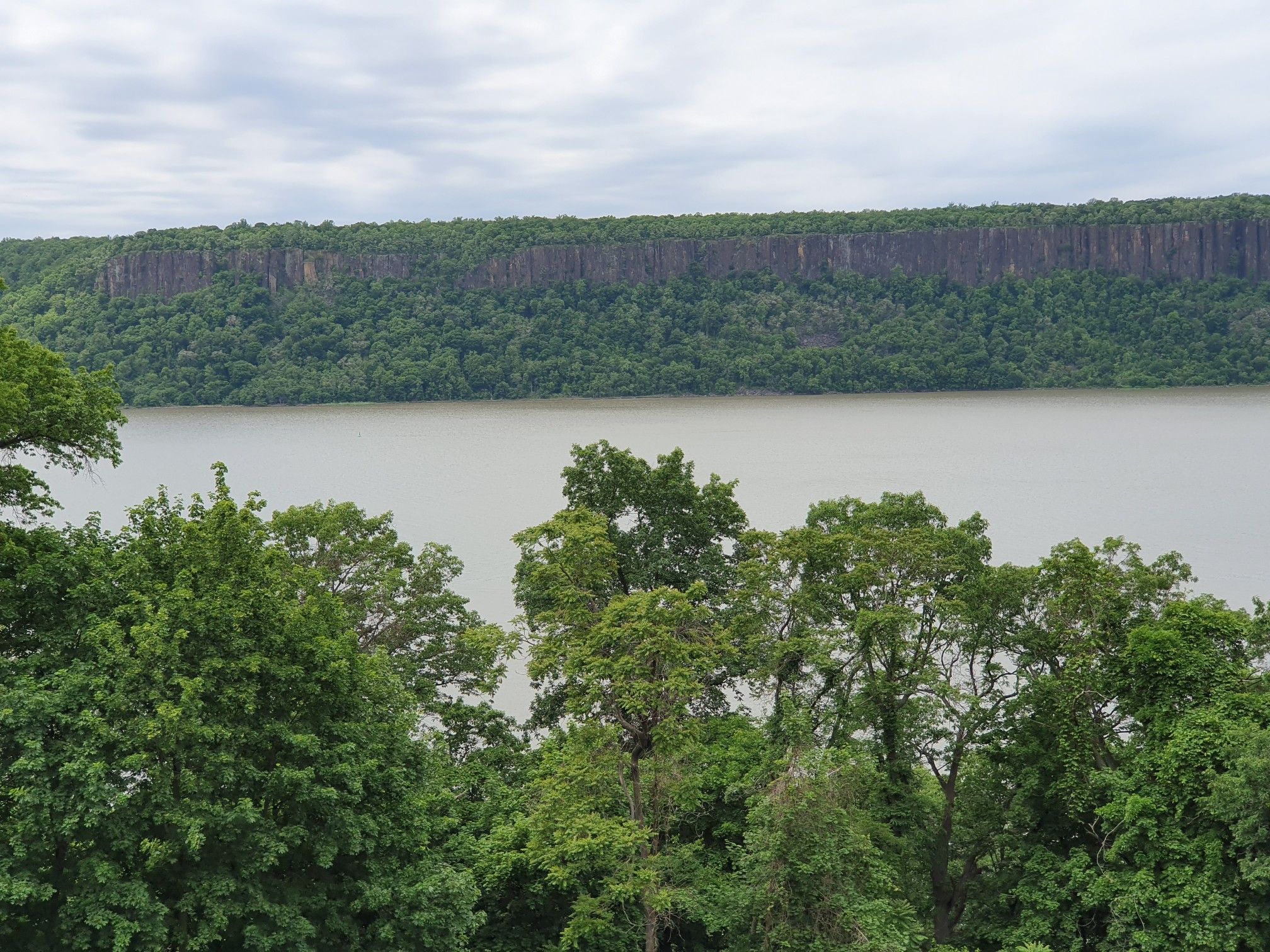 Wide river with distant forested cliff, green trees in foreground, overcast sky. - Untermyer Park Loop