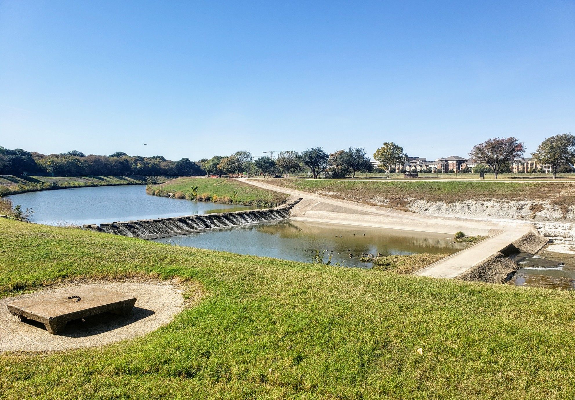 Sunny view of a river with grassy banks, a small dam, and distant buildings. - Trinity Trails