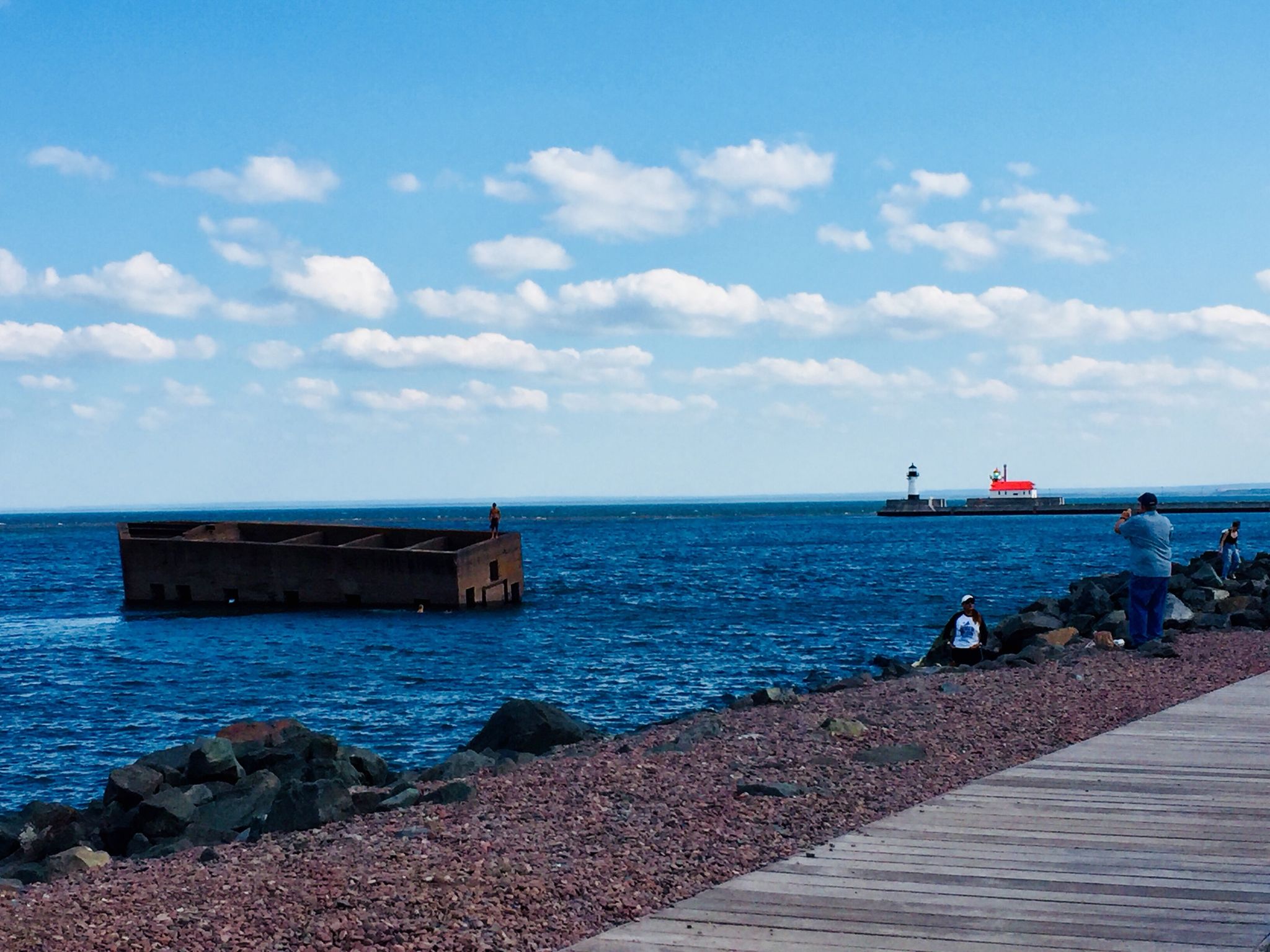 People on a boardwalk and shore near a floating structure and lighthouse. - Canal Park – Duluth Lakewalk (Canal Park) loop from Duluth Union Depot