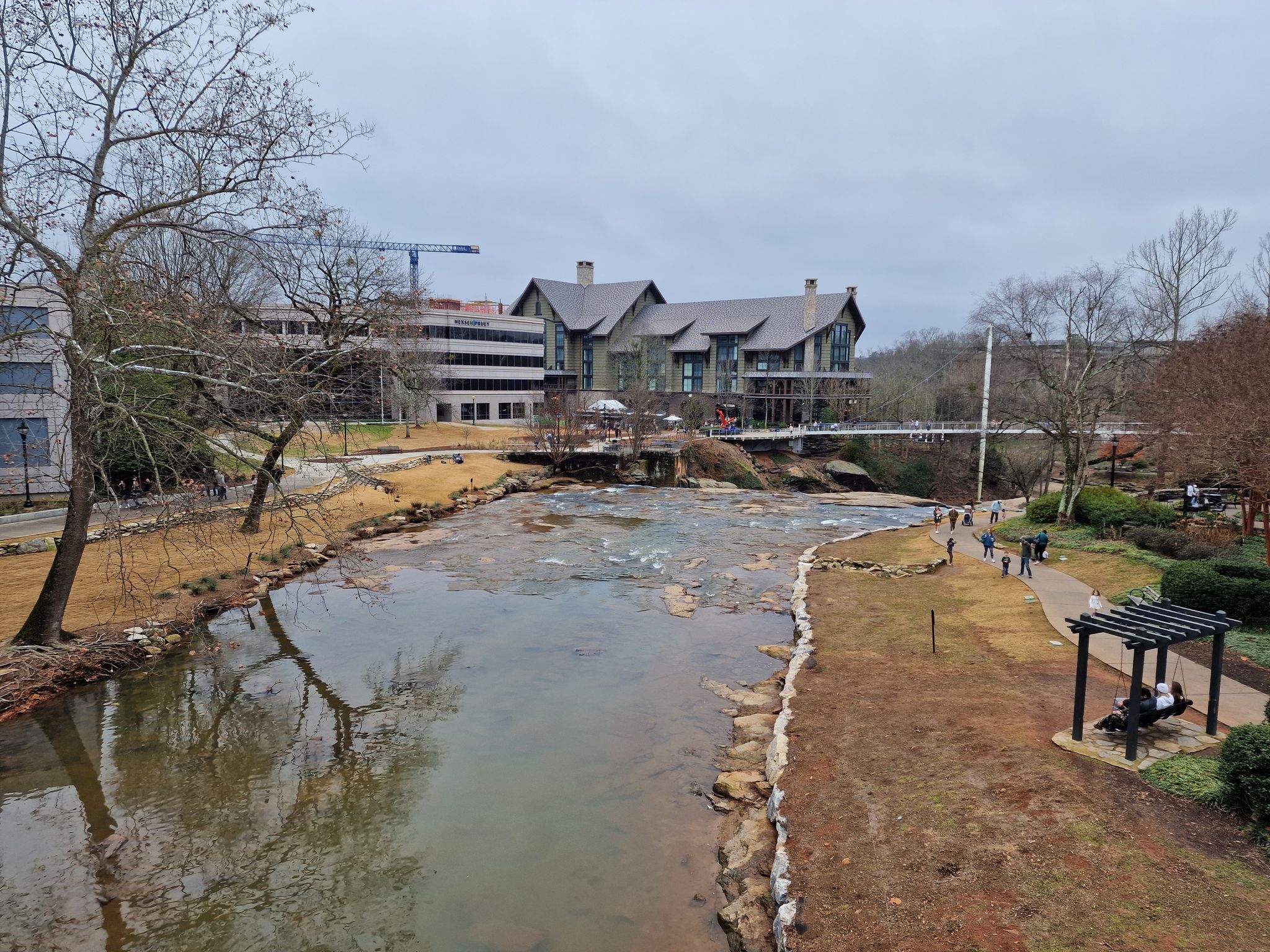 Overcast view of a river flowing past buildings and bare trees, with people walking. - Prisma Health Swamp Rabbit Trail