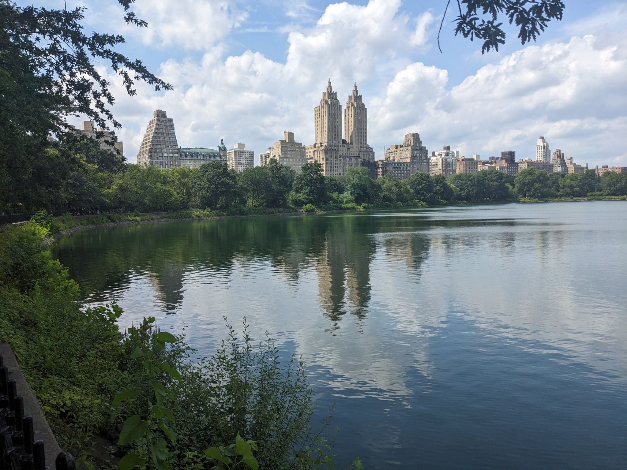 A wide lake reflects a distant city skyline under a bright, cloudy sky. - Manhattan and Brooklyn Waterfront Greenways Loop