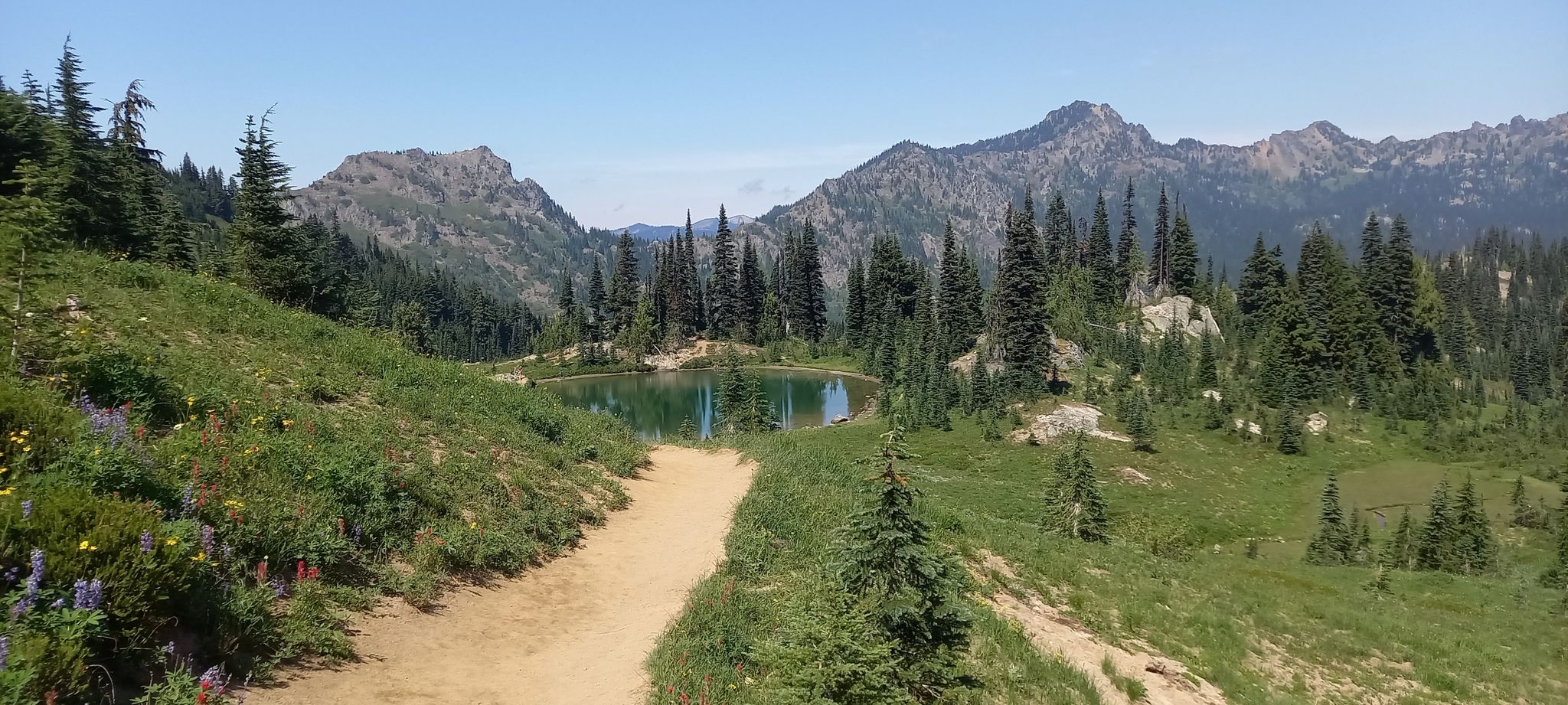 Dirt path leading to a small, calm lake surrounded by trees and mountains. - Dewey Lake Trail