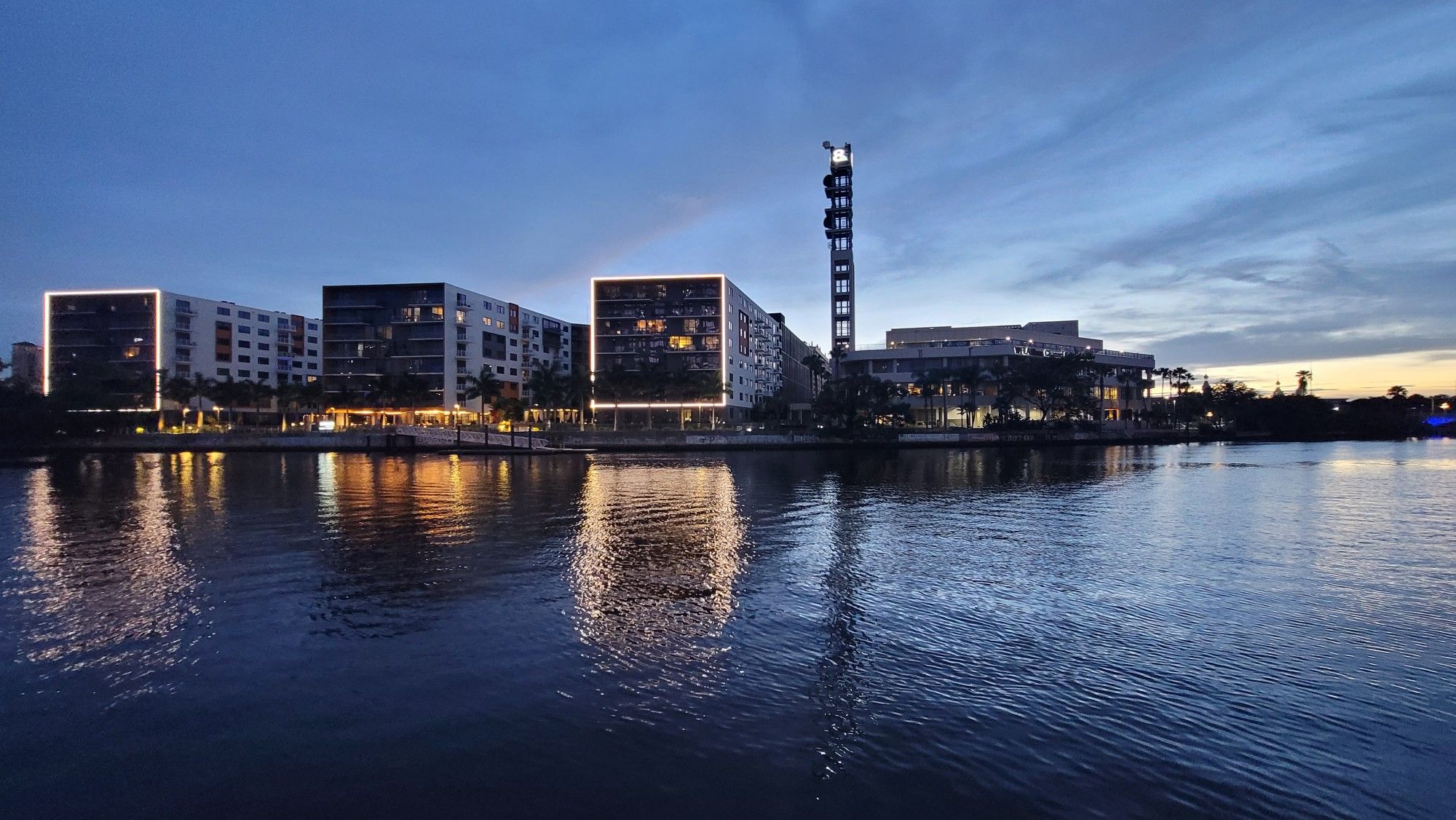 Illuminated buildings and a tall structure reflecting in dark water at dusk. - Tampa Riverwalk