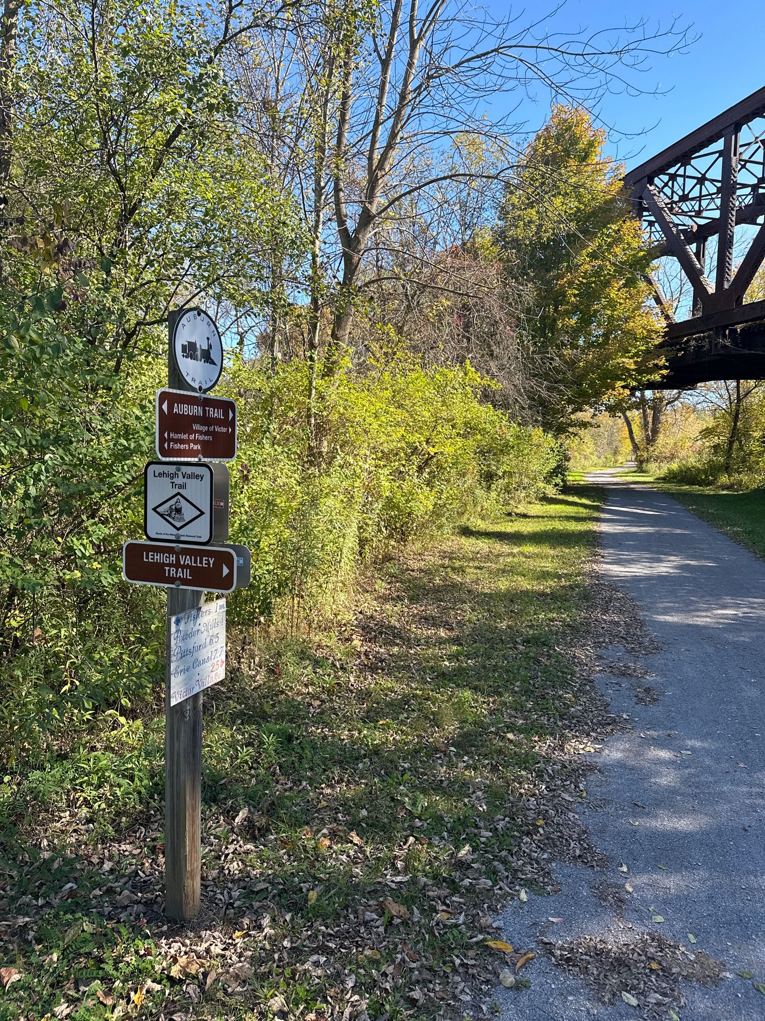 Trail signs beside a gravel path with a large metal bridge in autumn sunlight. - Old rock building – Victor trailhead loop from Victor
